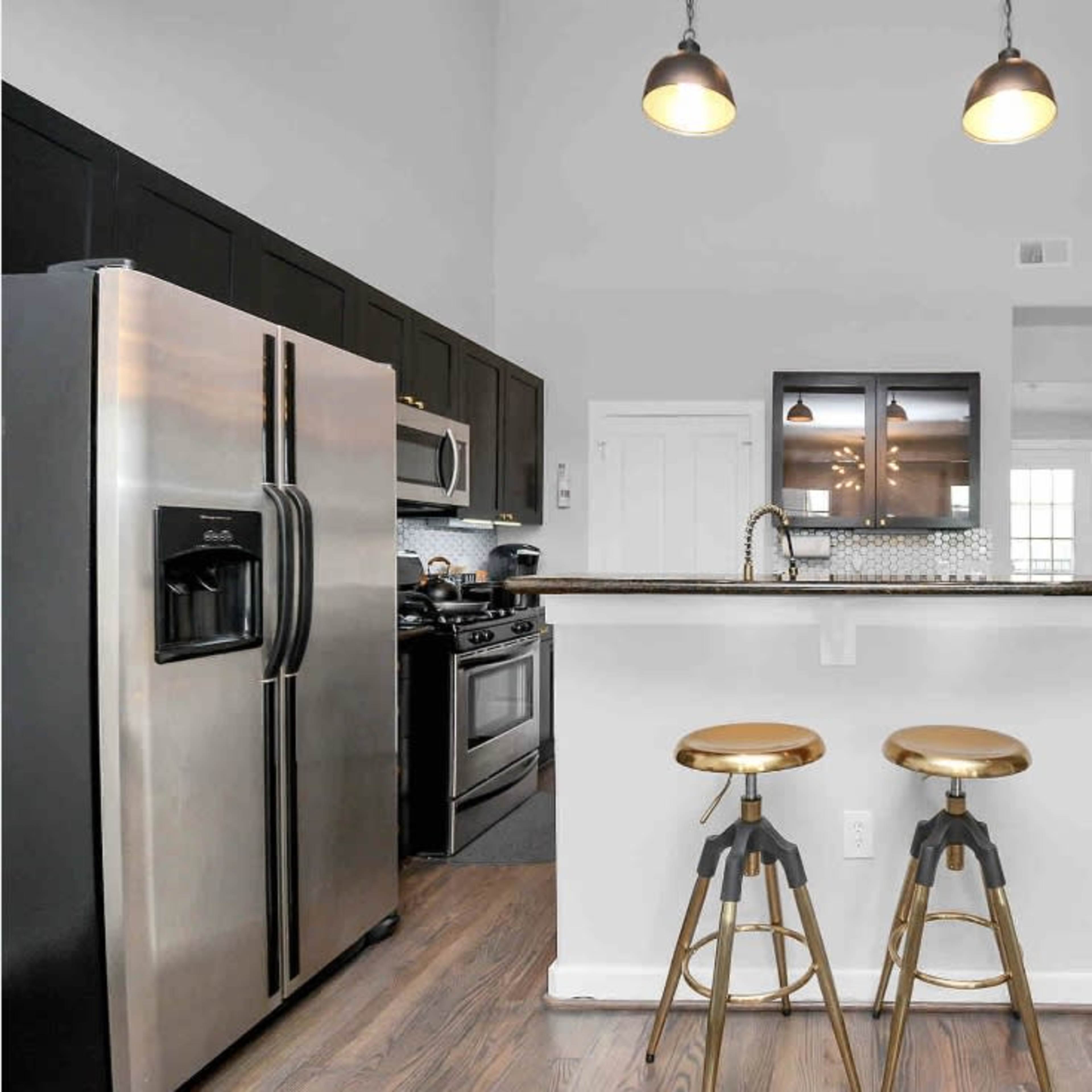 A modern kitchen features black cabinetry, stainless steel appliances, a central island with two stools, and a staircase in the background.