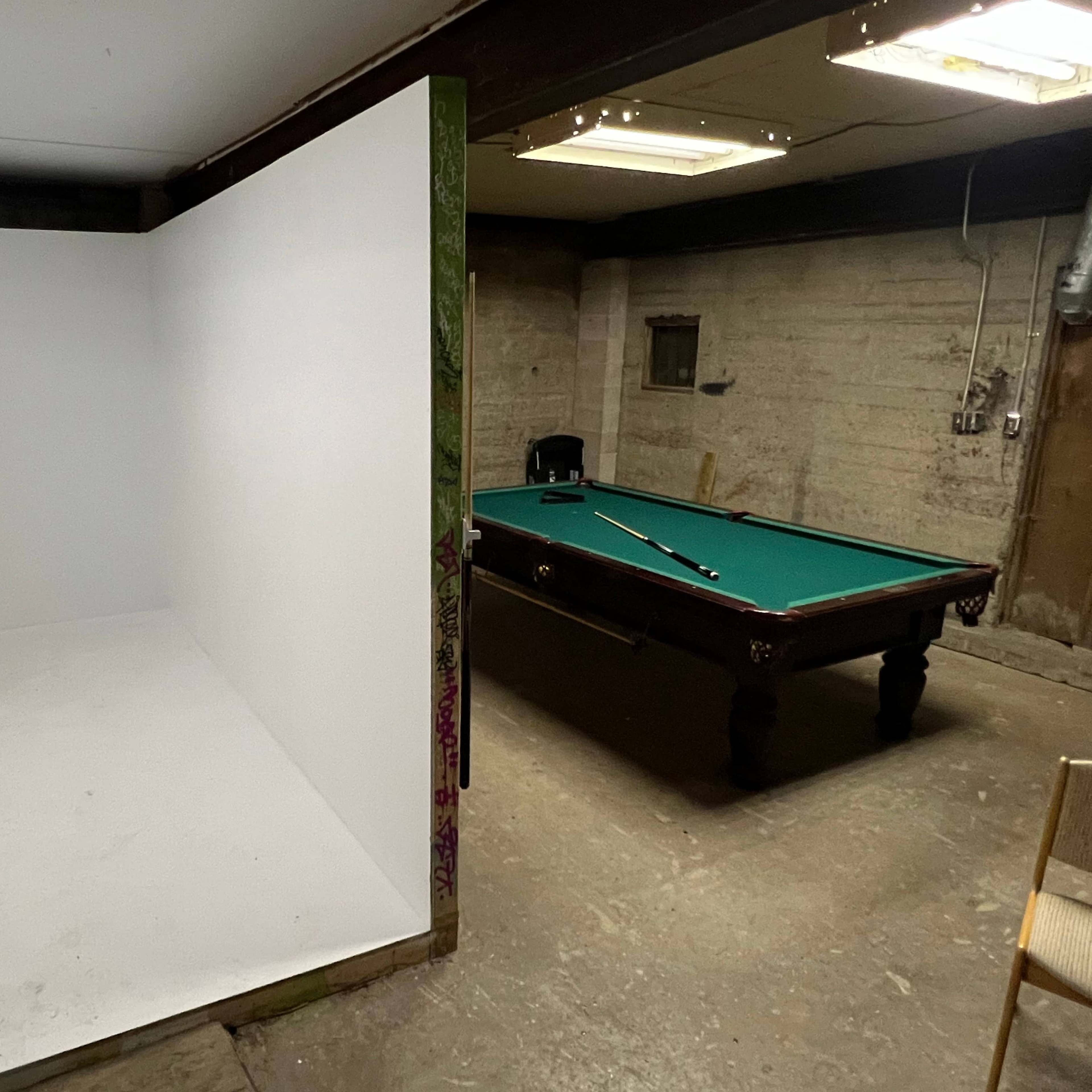 A bare white room is located adjacent to a pool table in a dimly lit basement.