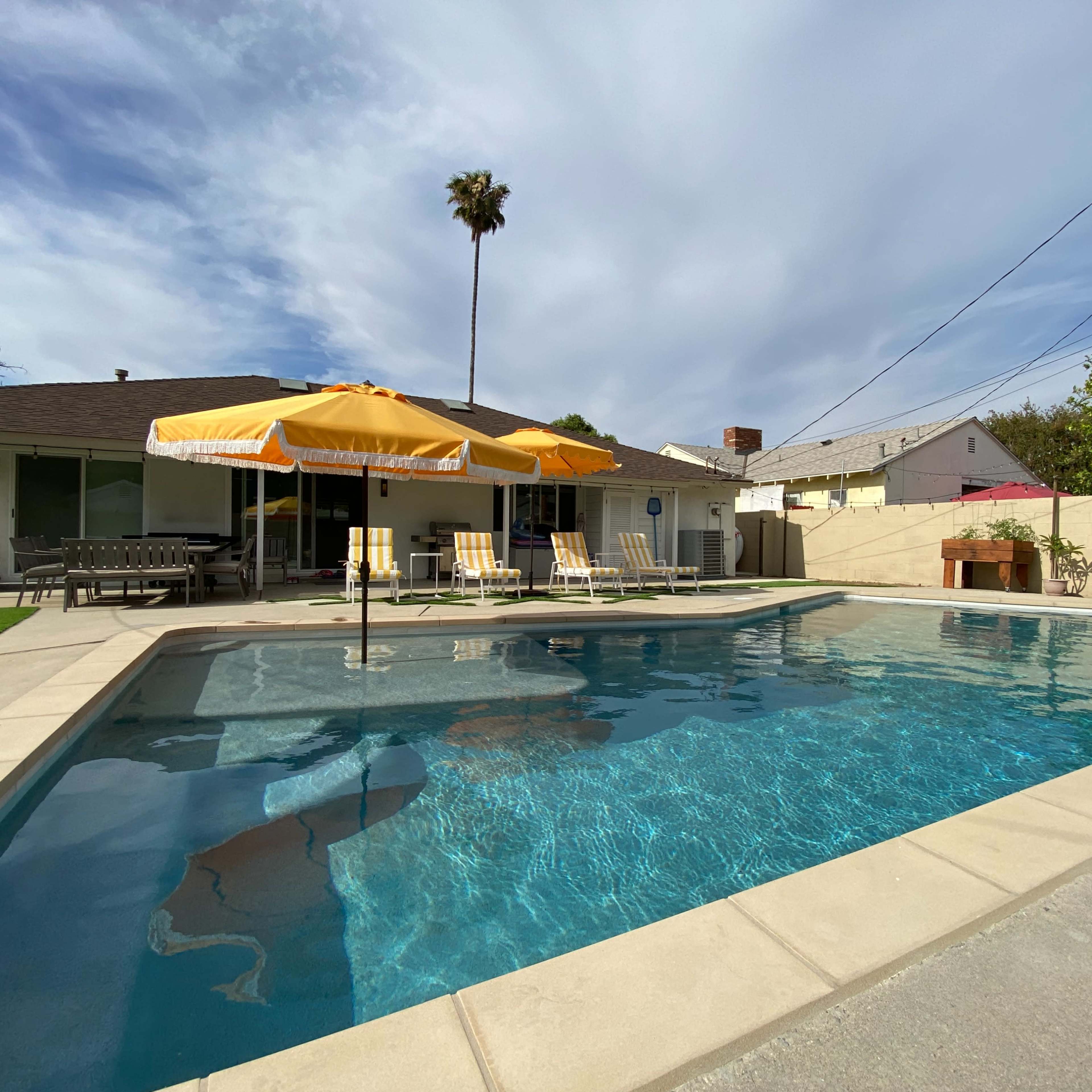 A rectangular swimming pool surrounded by lounge chairs and two yellow umbrellas beside a single-story house with a palm tree in the background.