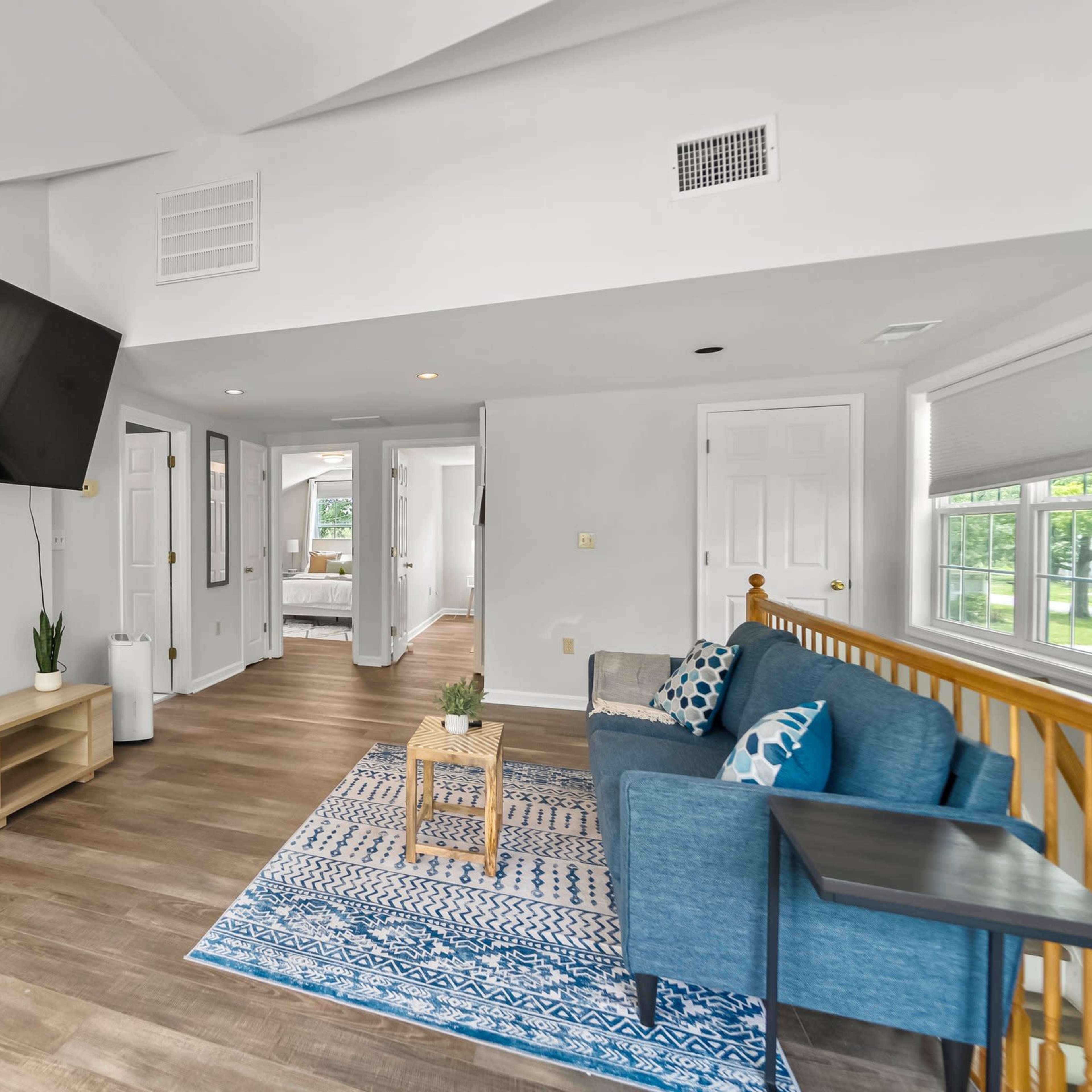 A bright, modern living space featuring a wall-mounted television, a blue couch, and a wooden coffee table on a patterned rug.