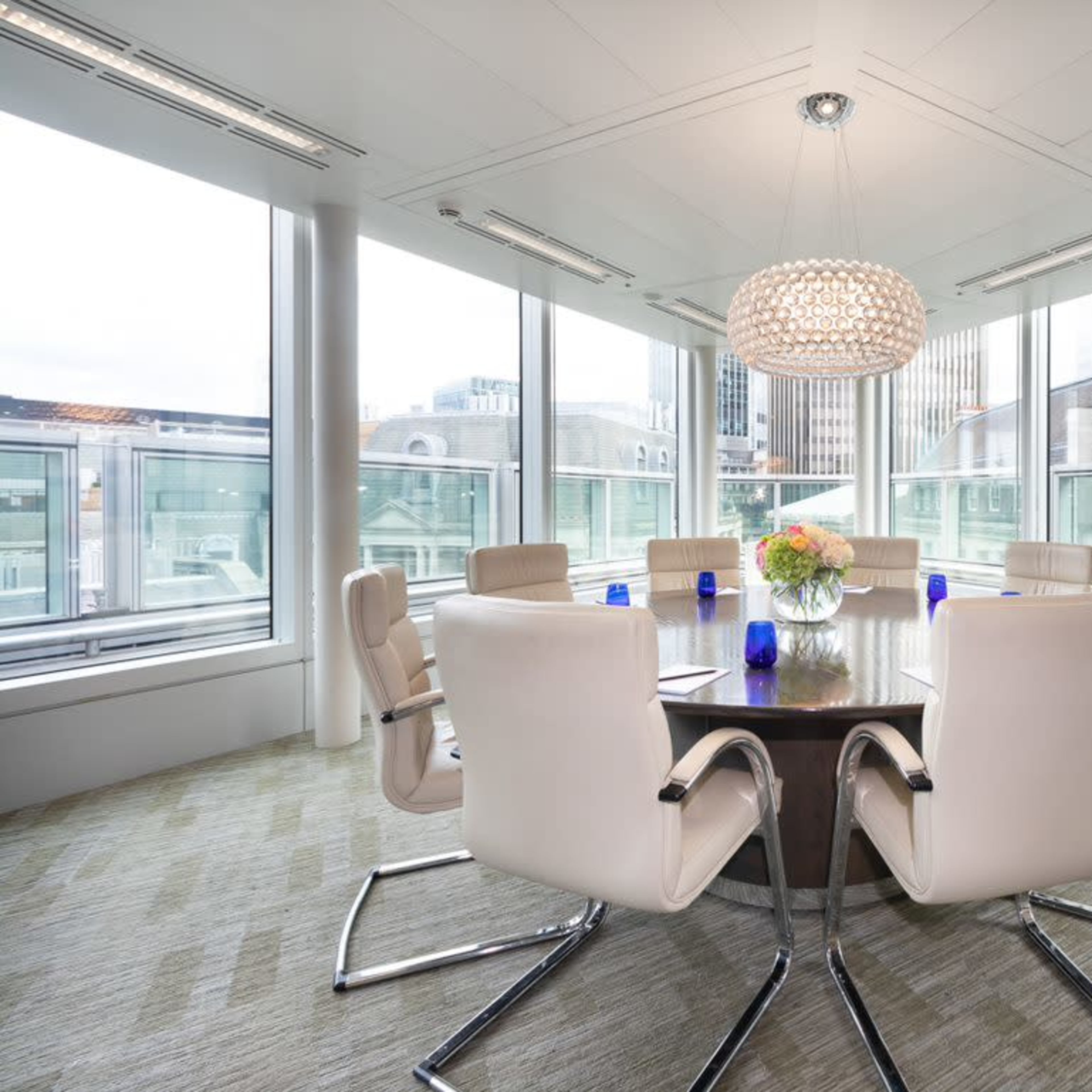 A conference room with a round table surrounded by white chairs and large windows offering a city view.