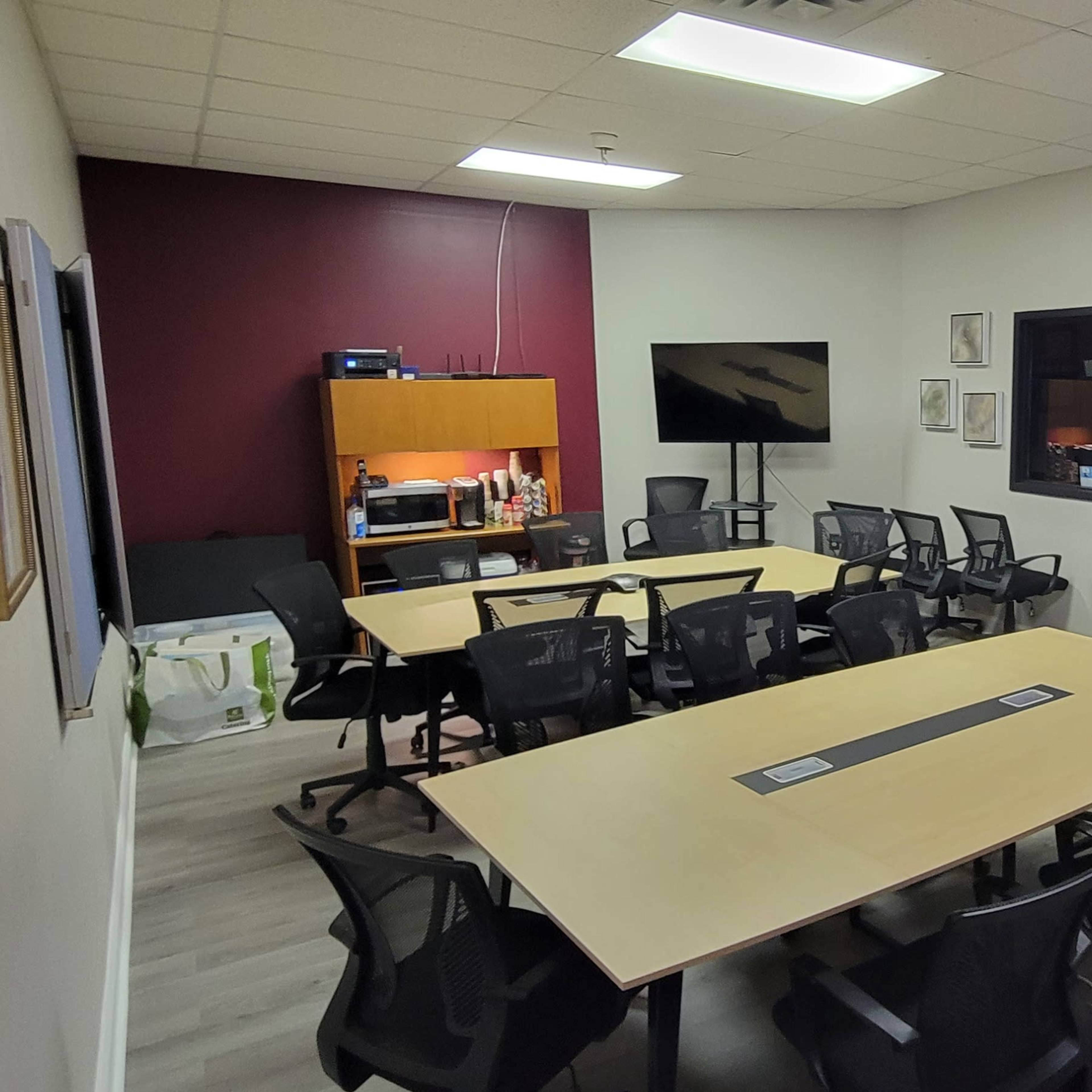 A conference room with a long table, multiple chairs, and a wall-mounted TV, with a large window displaying another room in the background.