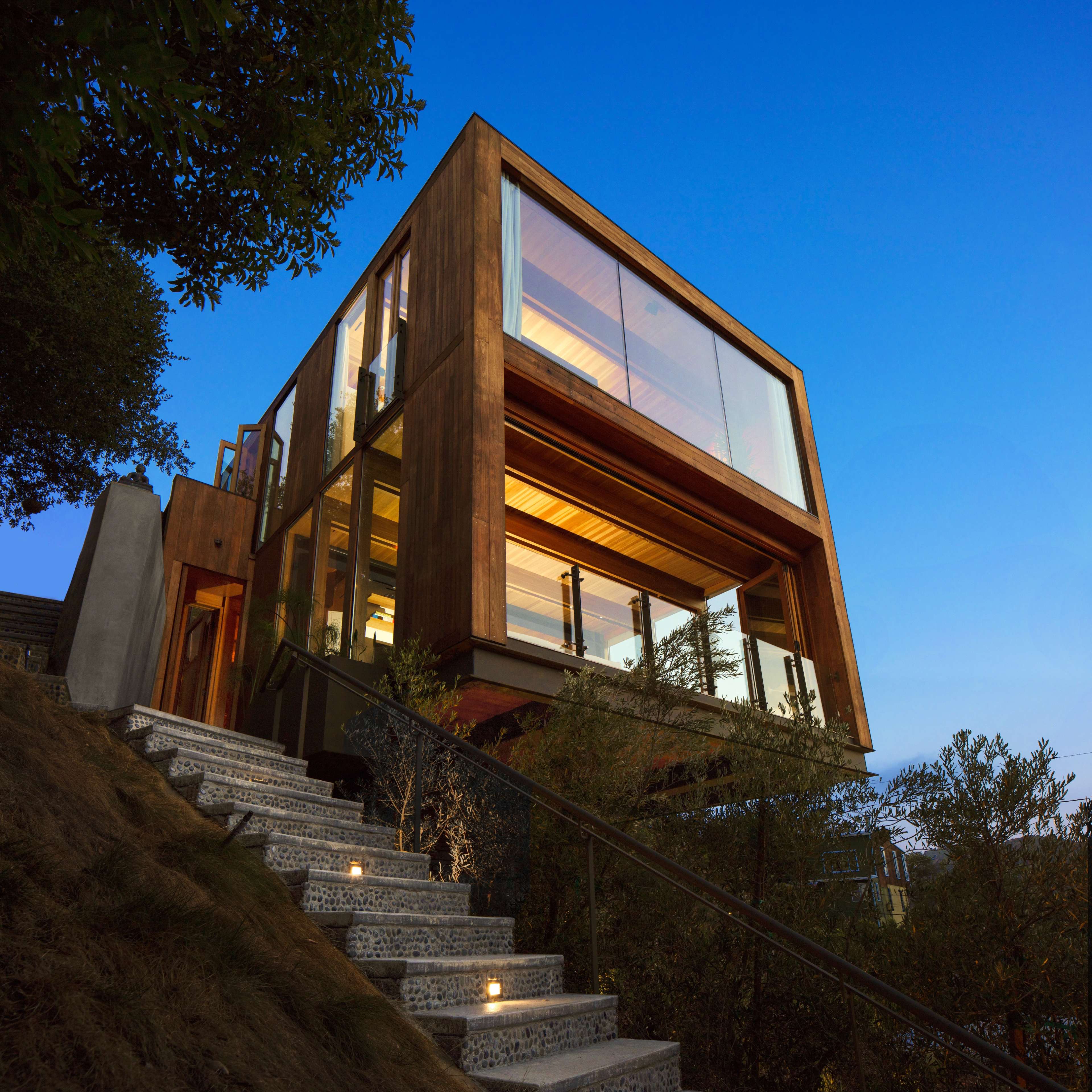 A modern wooden house with expansive glass windows is situated on a hillside, illuminated by outdoor lights along a stone pathway leading to its entrance.