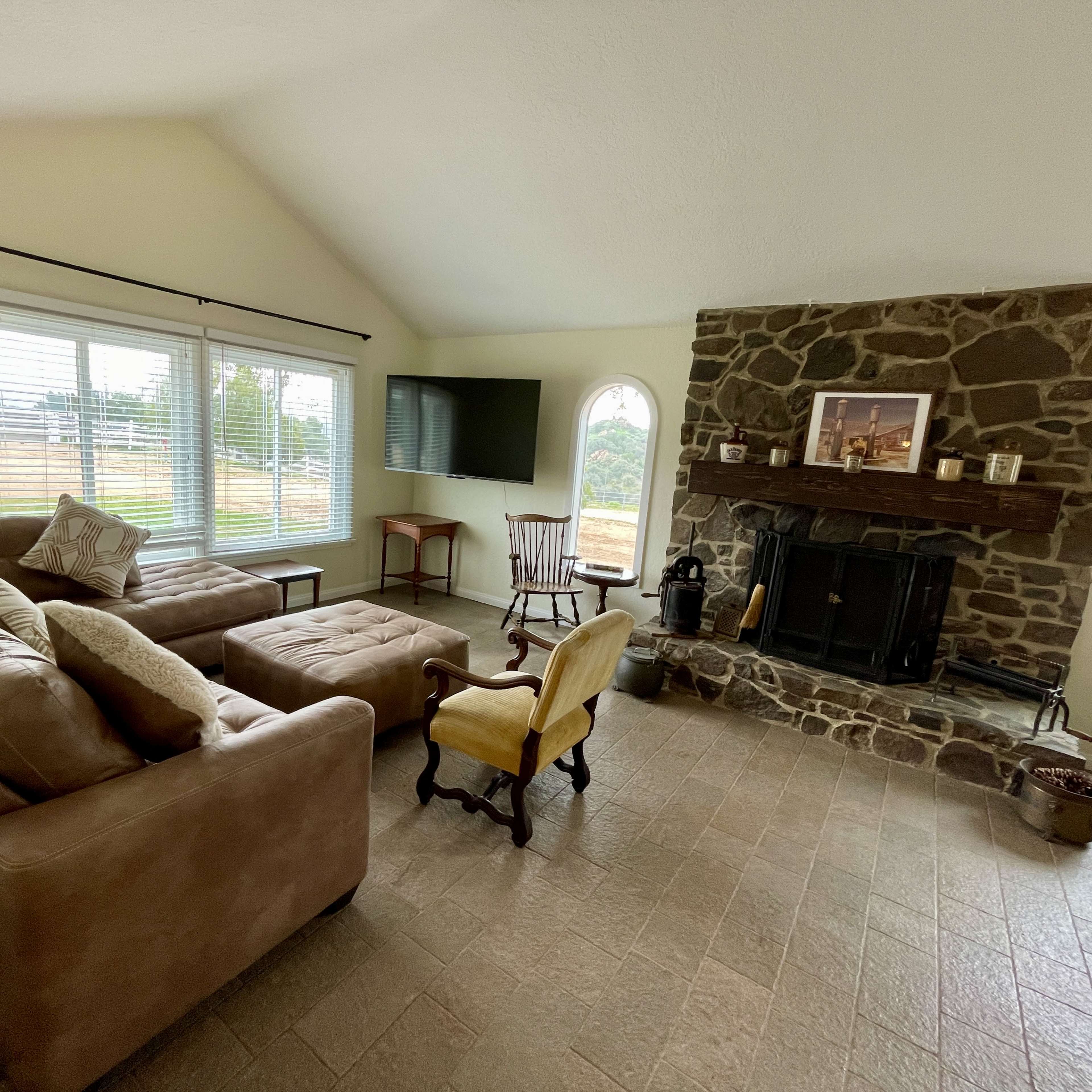 A living room features a stone fireplace with a wooden mantle, a sectional sofa, a yellow armchair, and large windows overlooking a yard.