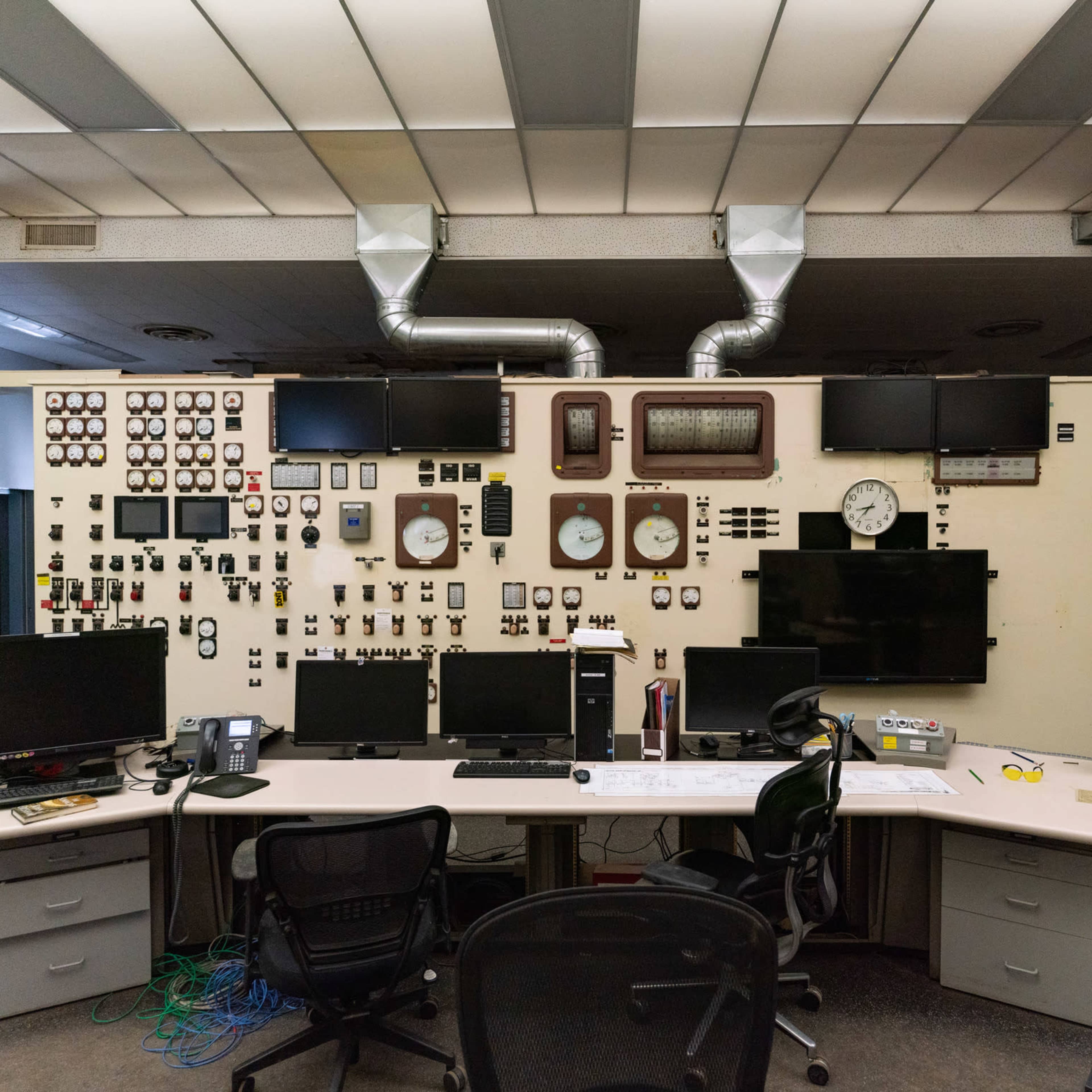 A control room with a large wall displaying various gauges, clocks, and monitors, along with a central desk equipped with multiple computer screens and chairs.
