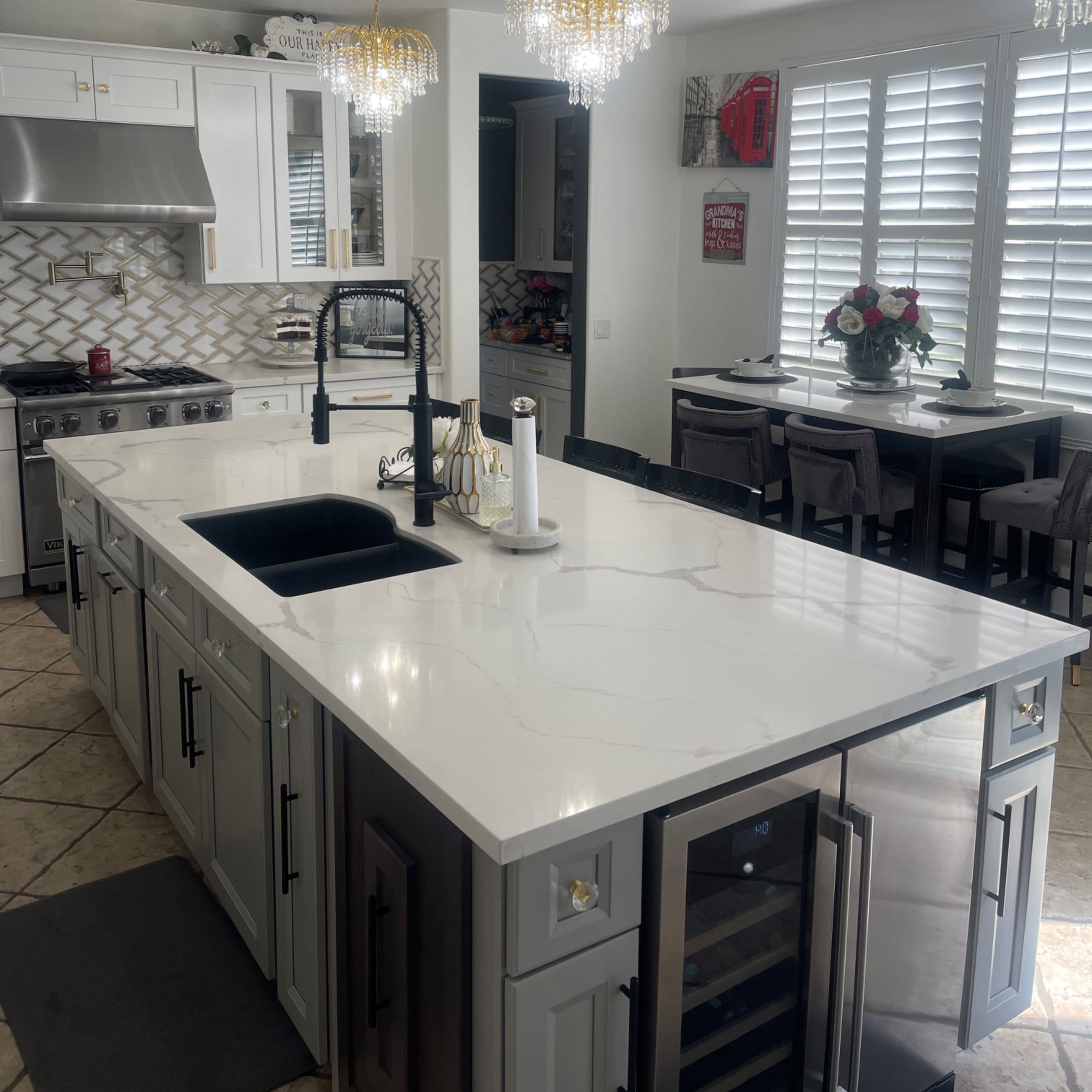 A modern kitchen with a central island, featuring a marble countertop, a sink, and stainless steel appliances, complemented by decorative lighting fixtures and a dining area in the background.