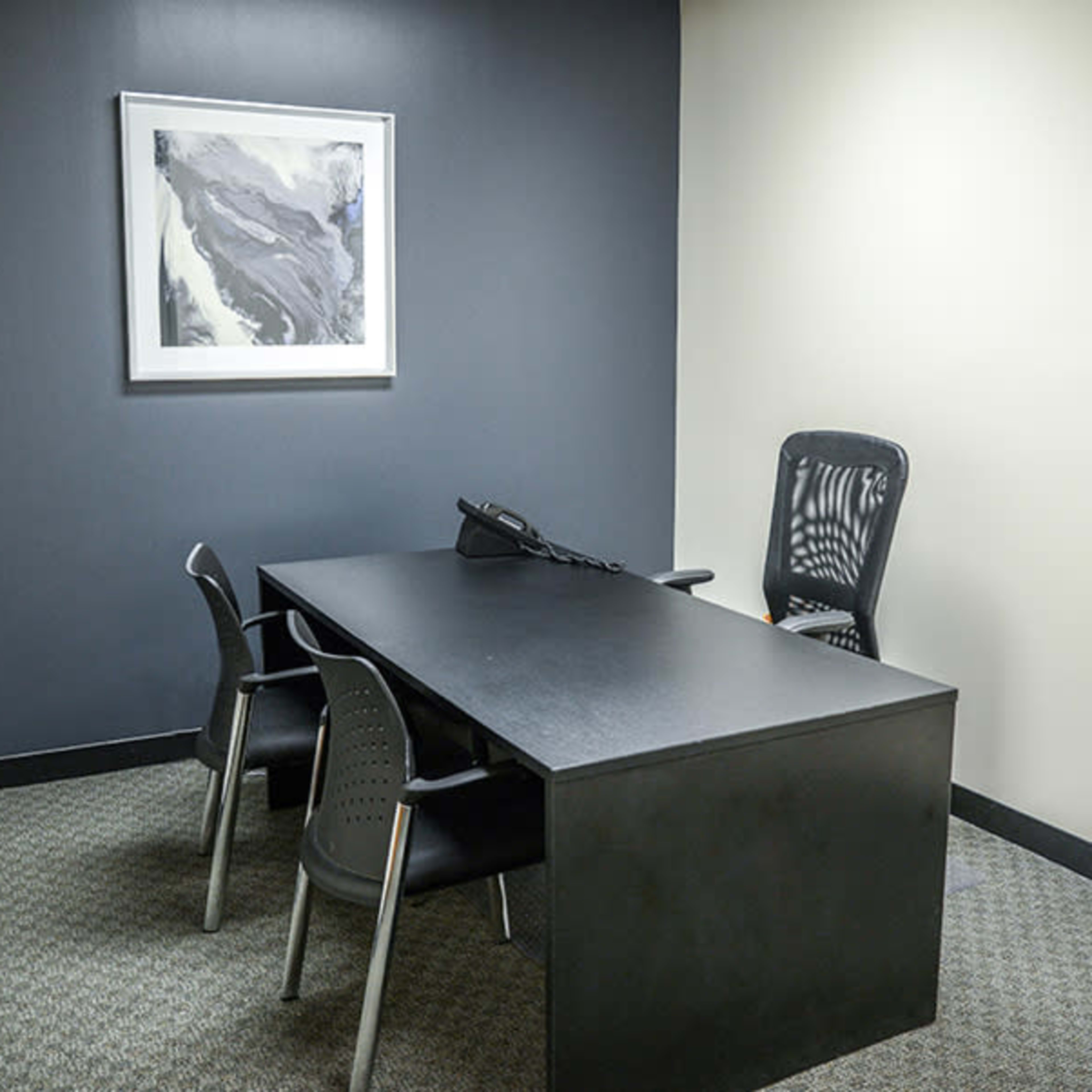 A small office space with a black desk, three chairs, a telephone, and a piece of artwork on the wall.