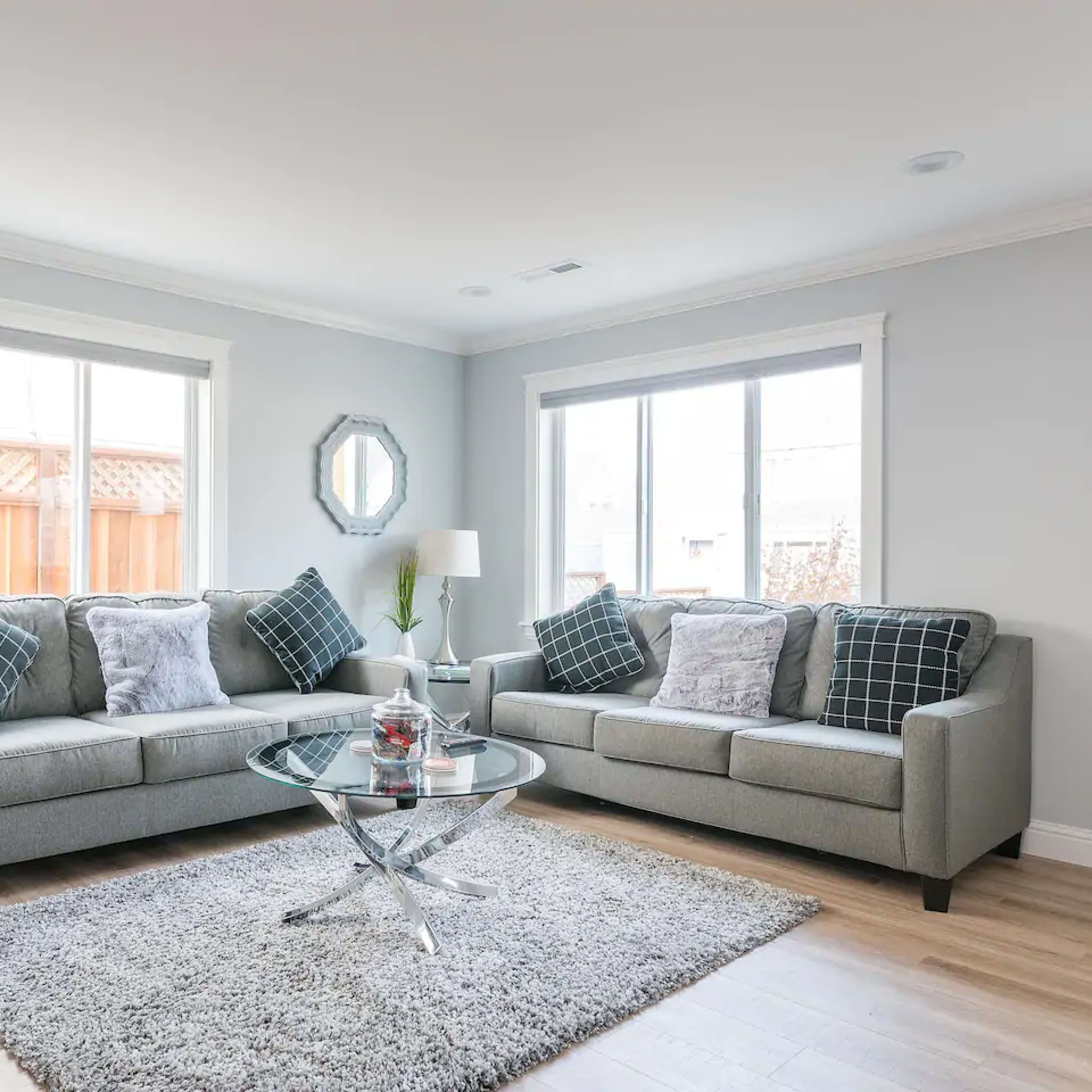 A modern living room features two gray sofas with plaid pillows, a circular glass coffee table, and large windows allowing natural light.