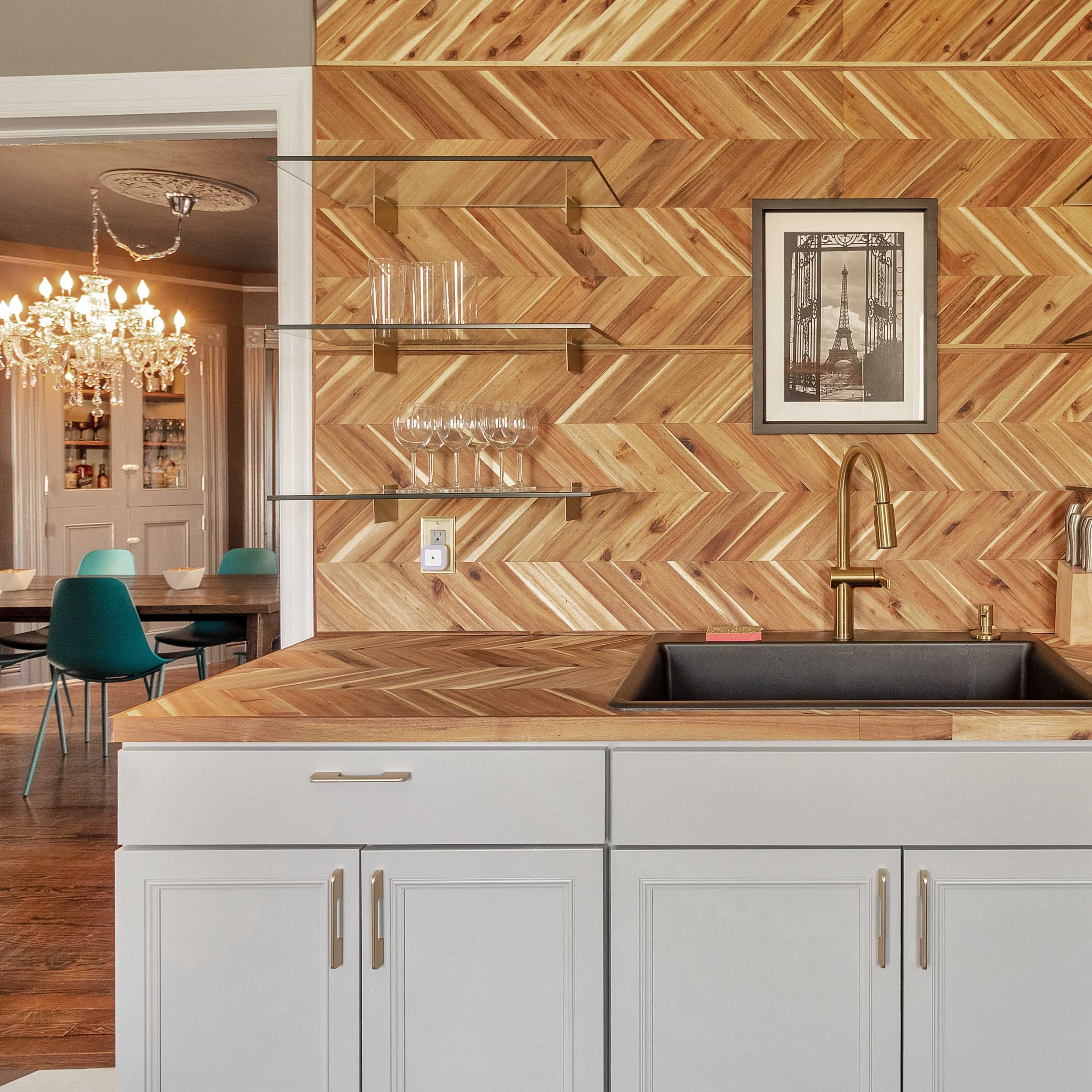 A kitchen with a herringbone-patterned wooden wall, a gray cabinetry with a sink and a small fridge, and a dining area visible in the background.
