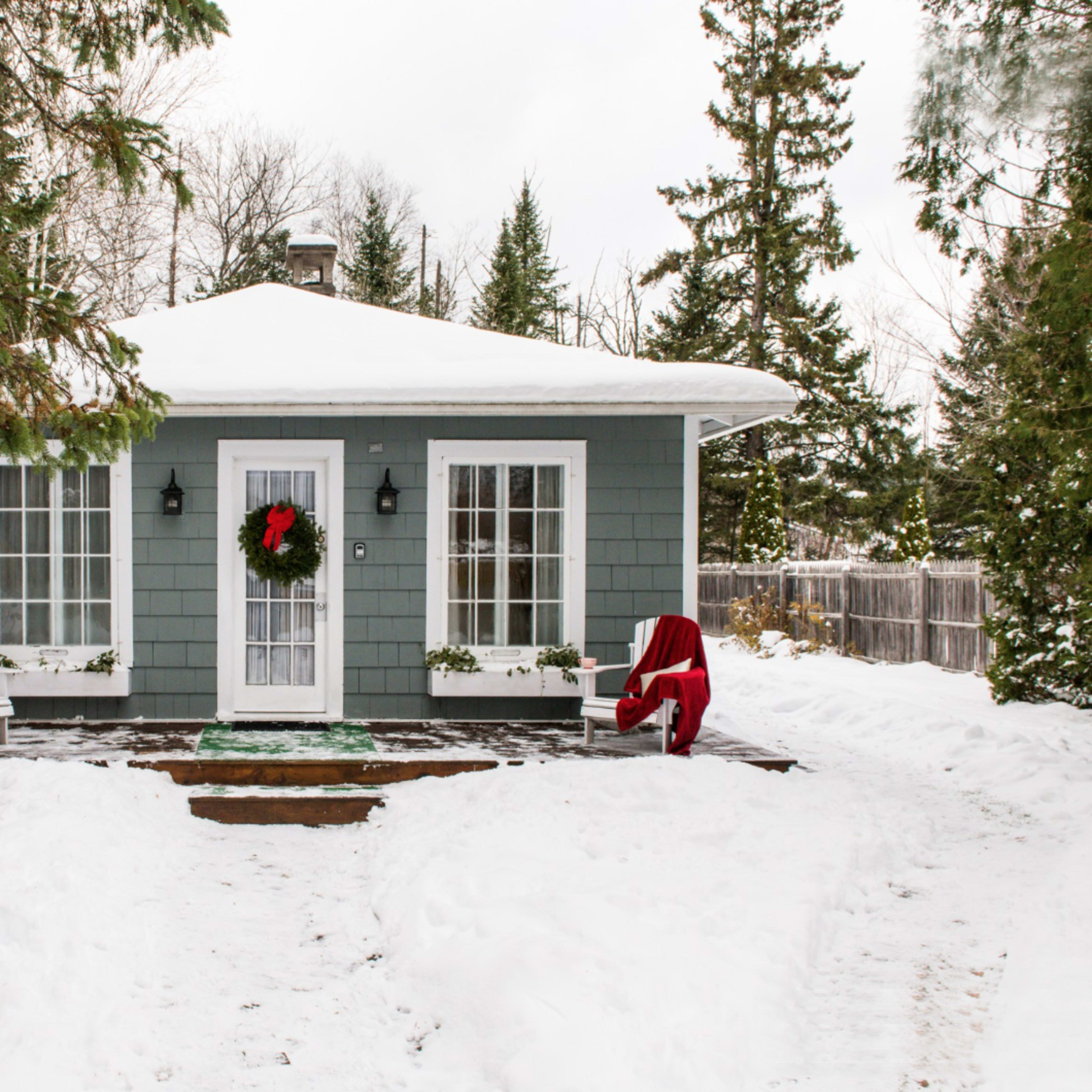 A small, snow-covered cottage with a green exterior features a festive wreath on the door and a red chair on the porch.