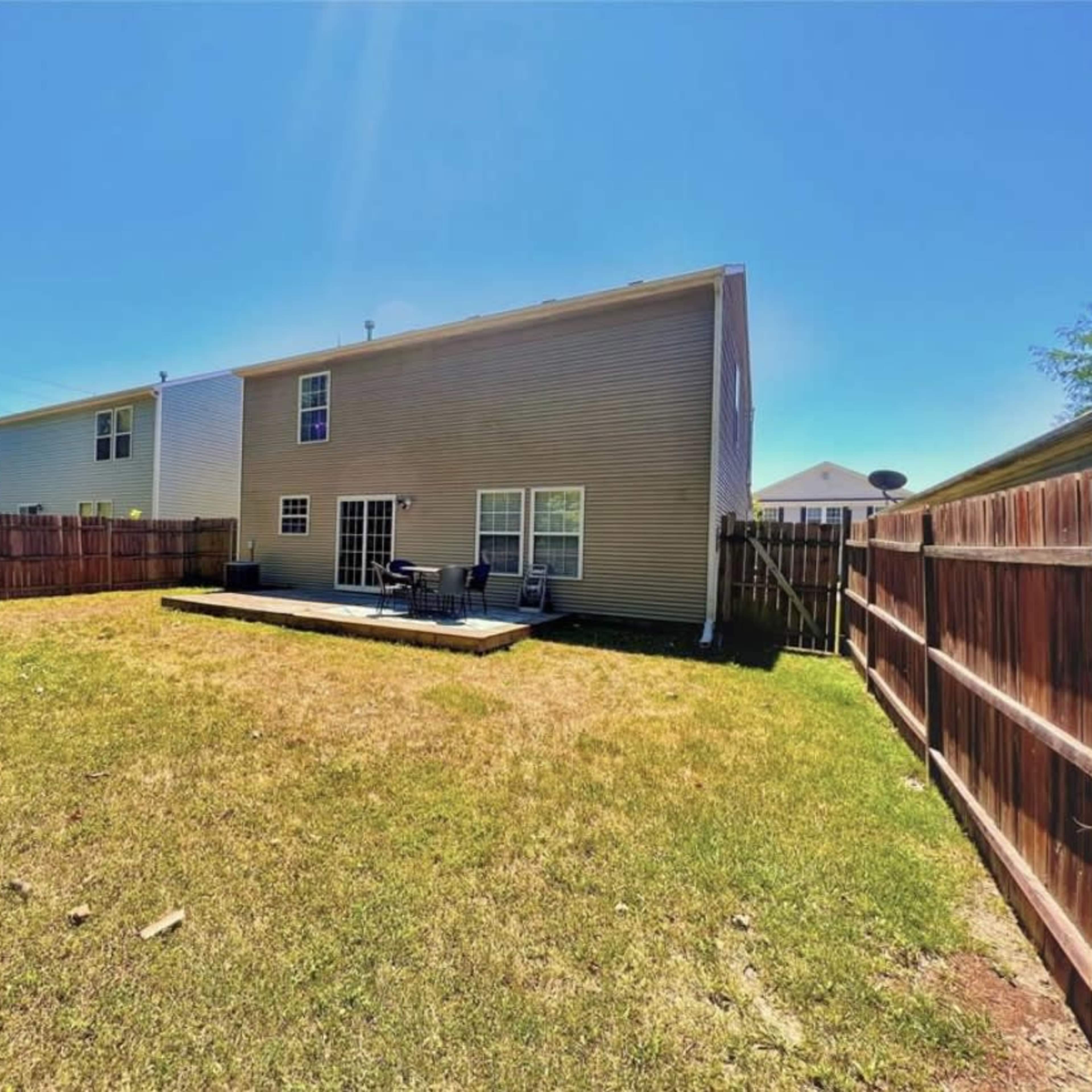 A backyard enclosed by a wooden fence, featuring a patch of grass and a small patio area with chairs.