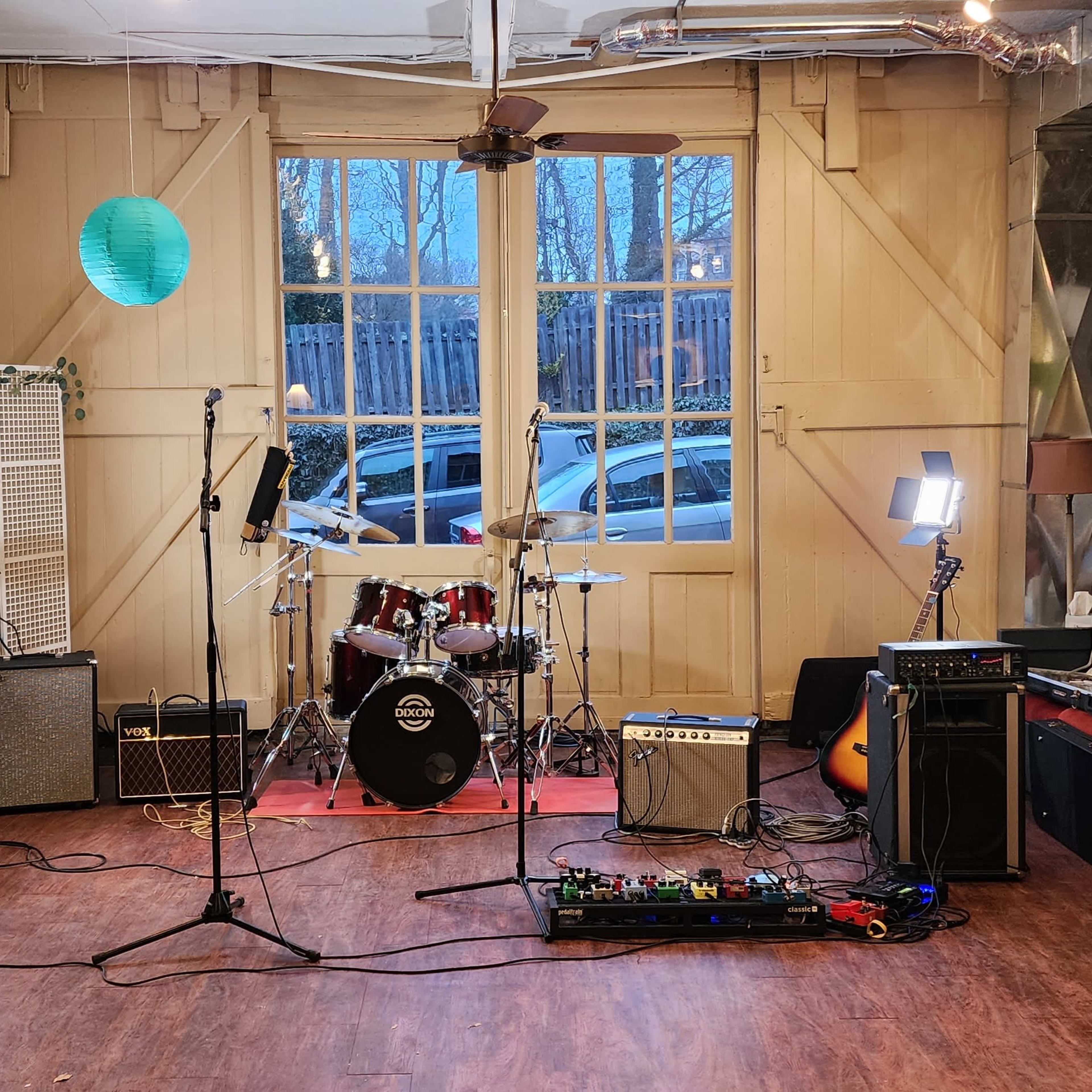 A music rehearsal space with a drum set, amplifiers, and various instruments arranged around a stage area near large windows.