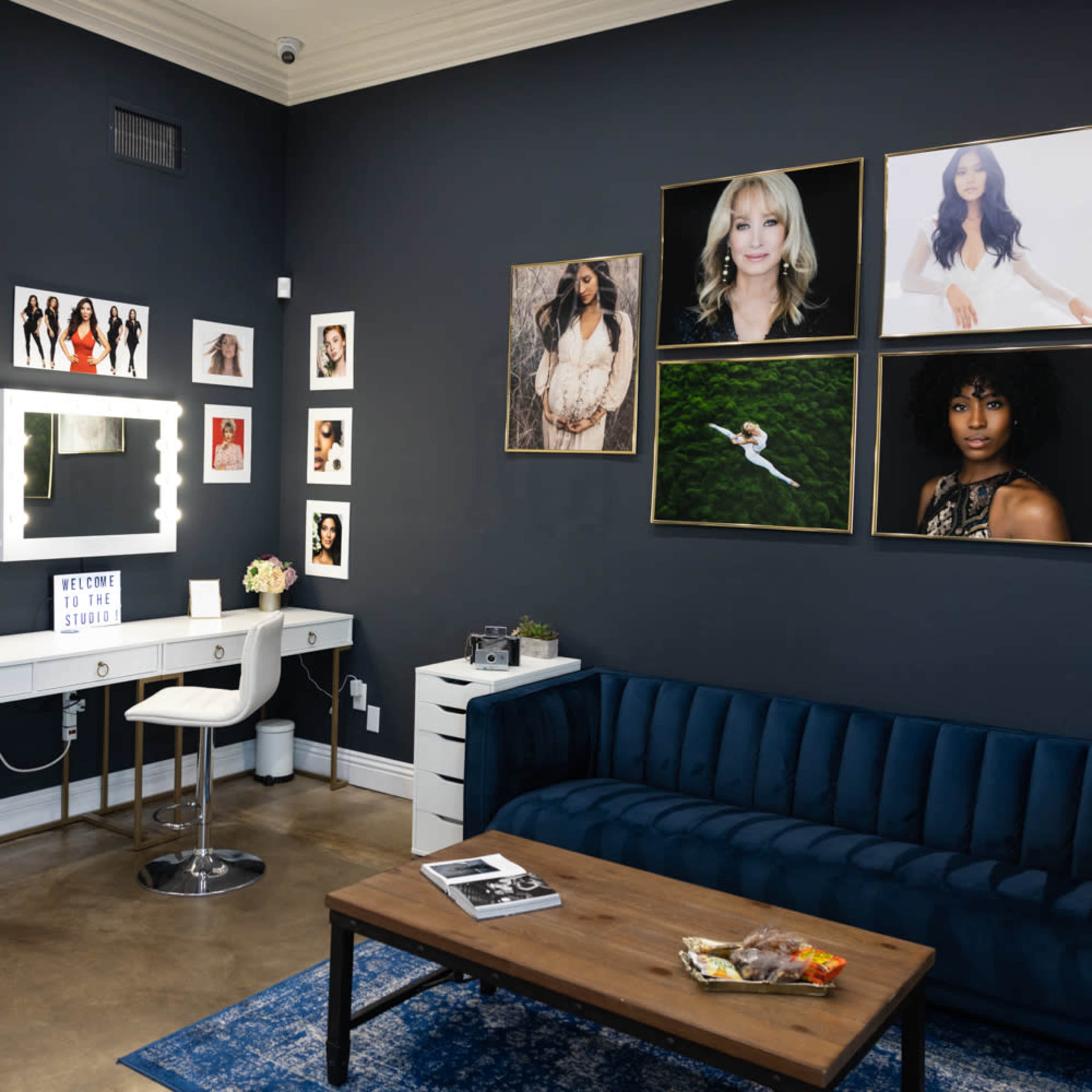 A modern beauty studio featuring a blue velvet sofa, a white vanity with a lighted mirror, and several framed portraits on the dark wall.