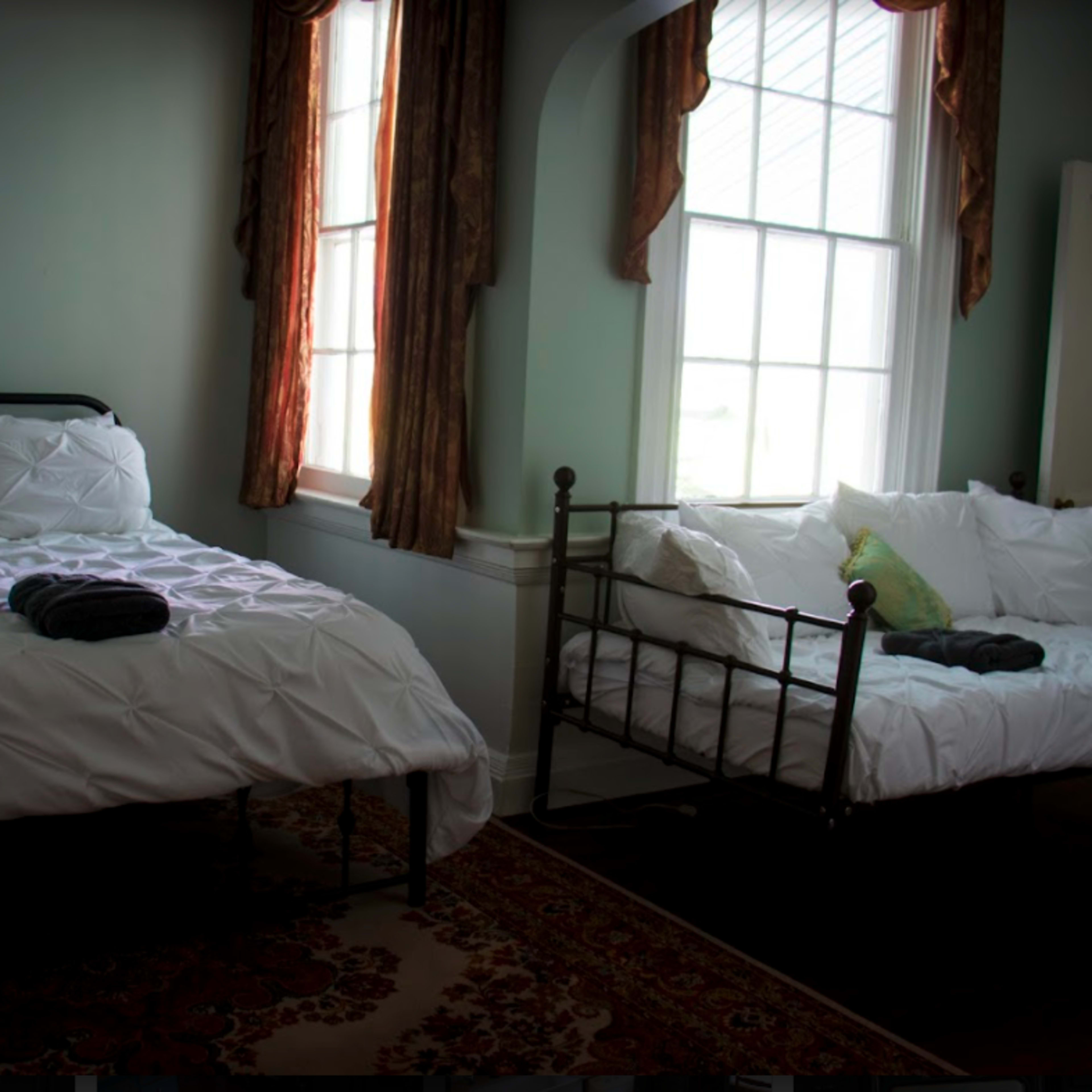 A bedroom with two beds, one a standard size and the other a smaller daybed, both dressed in white linens, with large windows letting in natural light.