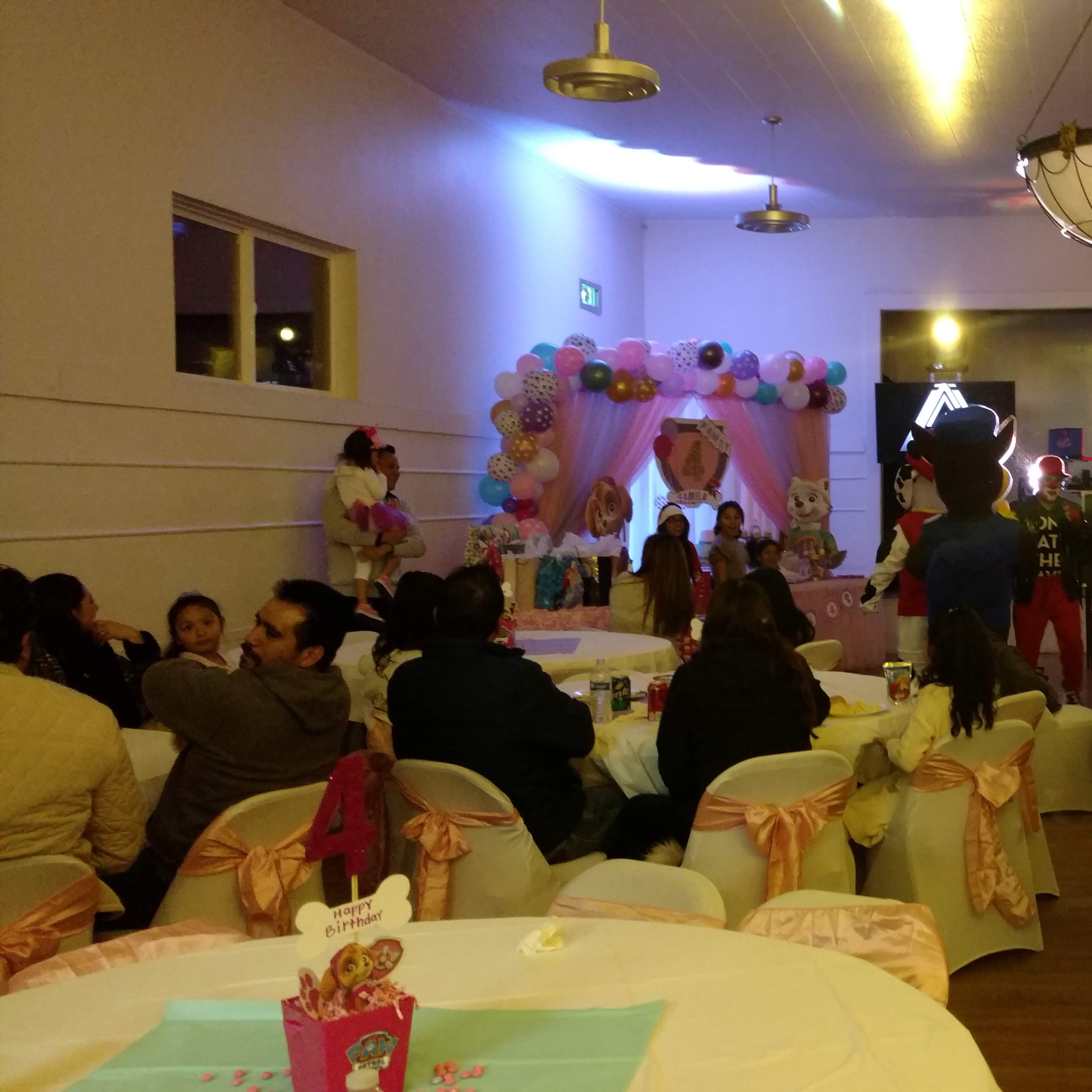 A party scene is set indoors, with tables adorned in pink and white decorations, featuring balloons and a stage with lighting effects in the background.