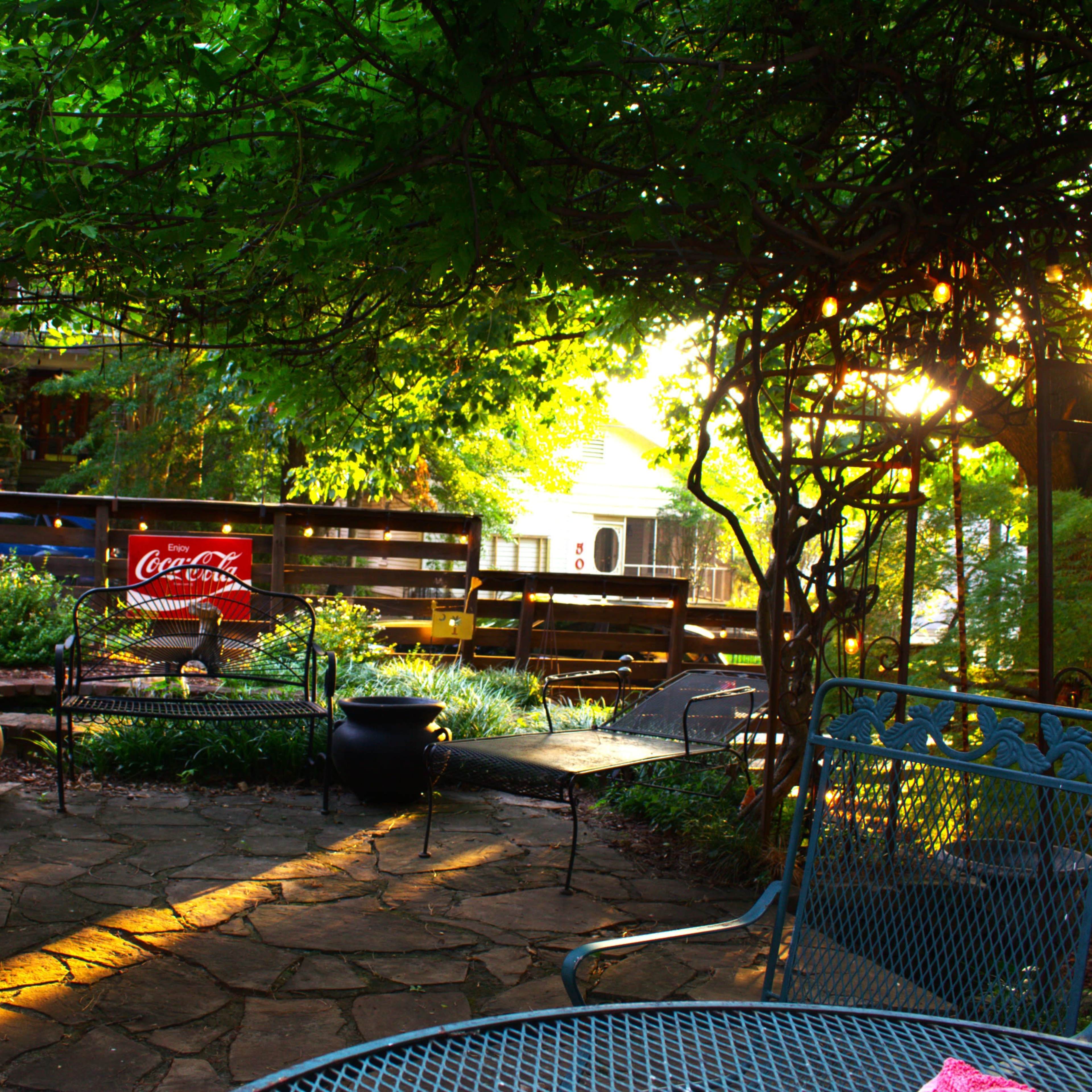 A patio area surrounded by greenery, featuring metal furniture and a Coca-Cola sign against a wooden fence, with sunlight filtering through the leaves.