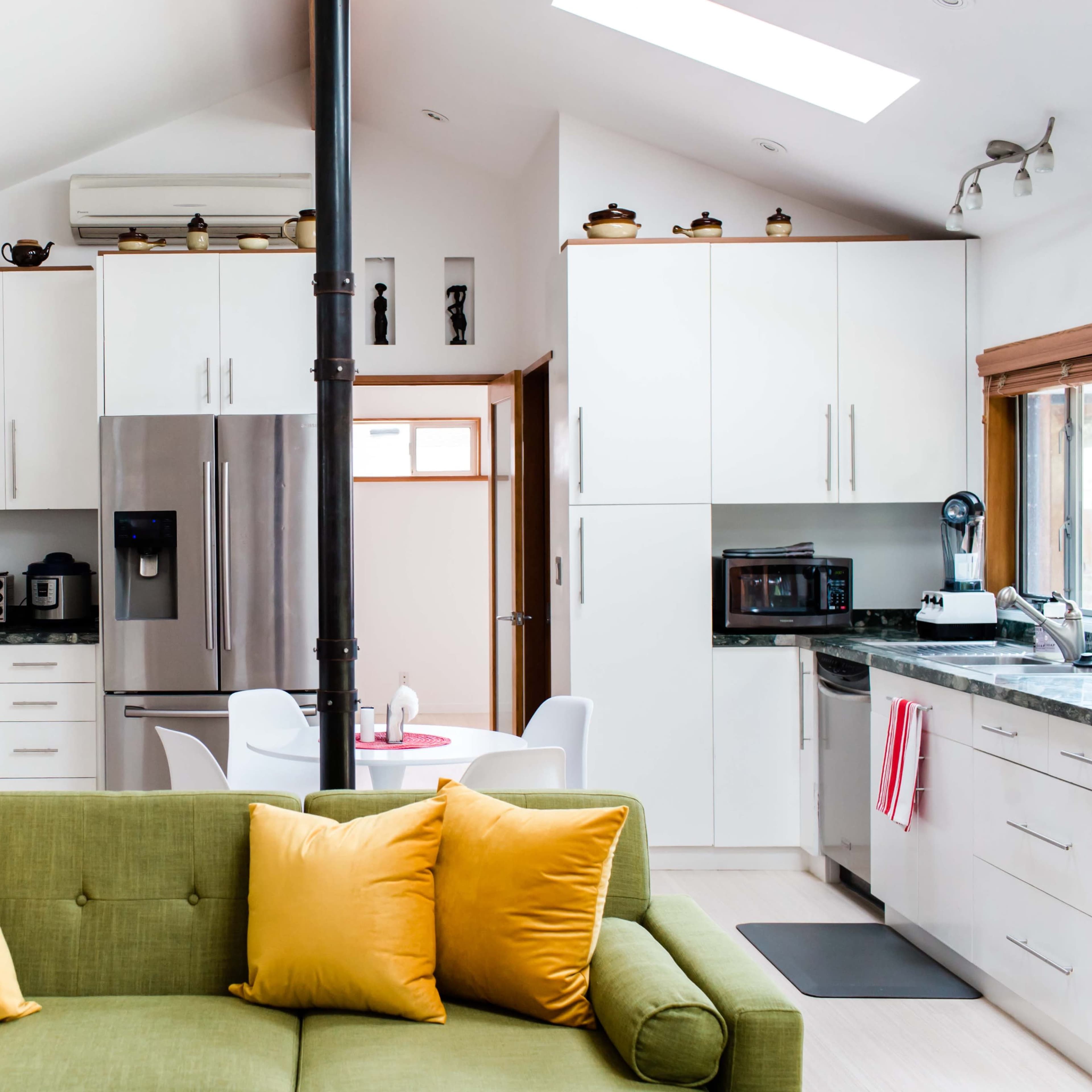 A modern kitchen features white cabinetry and a green sofa with yellow pillows, with a dining area visible in the background.