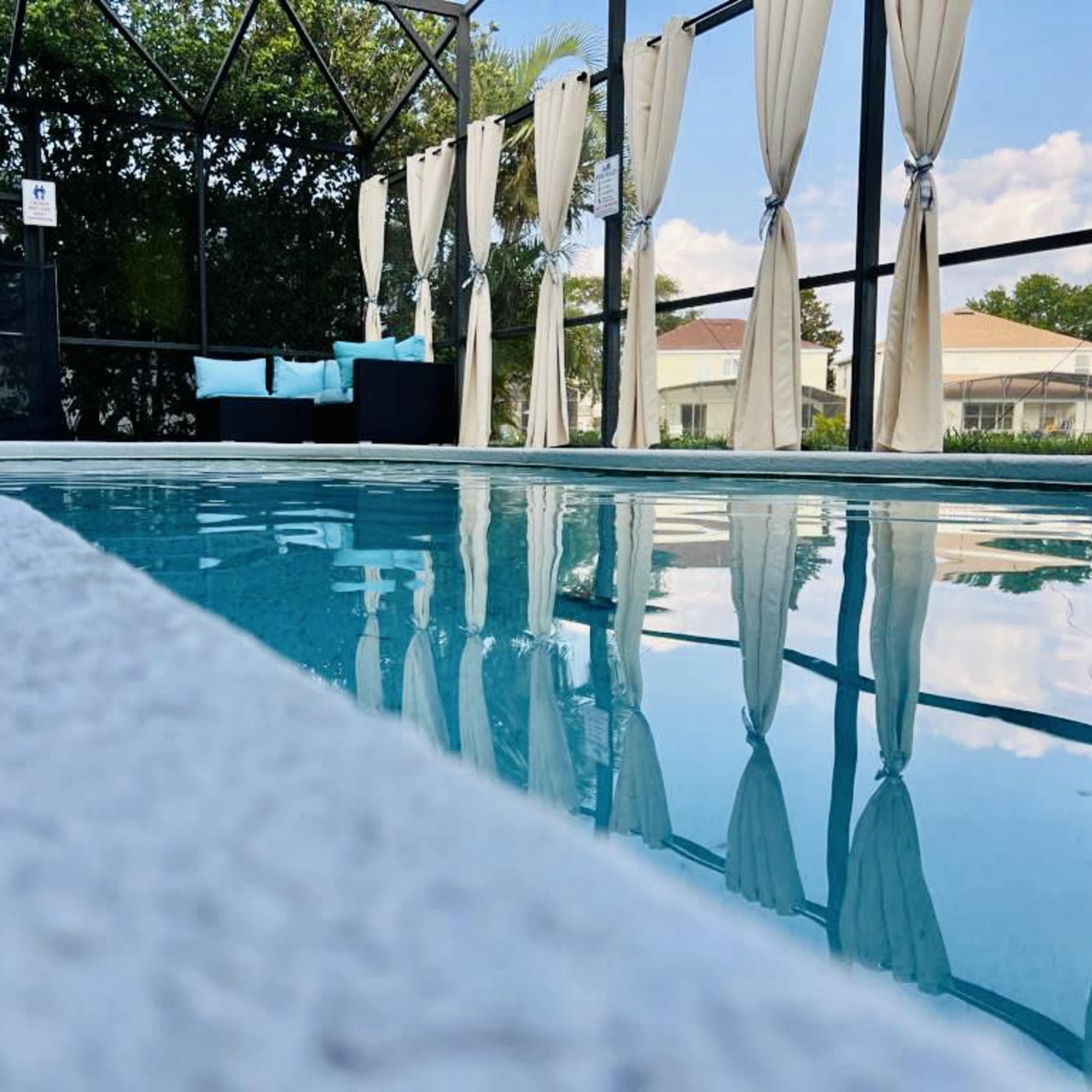 A tranquil swimming pool enclosed by a screened patio, with white curtains and a seating area in the background.