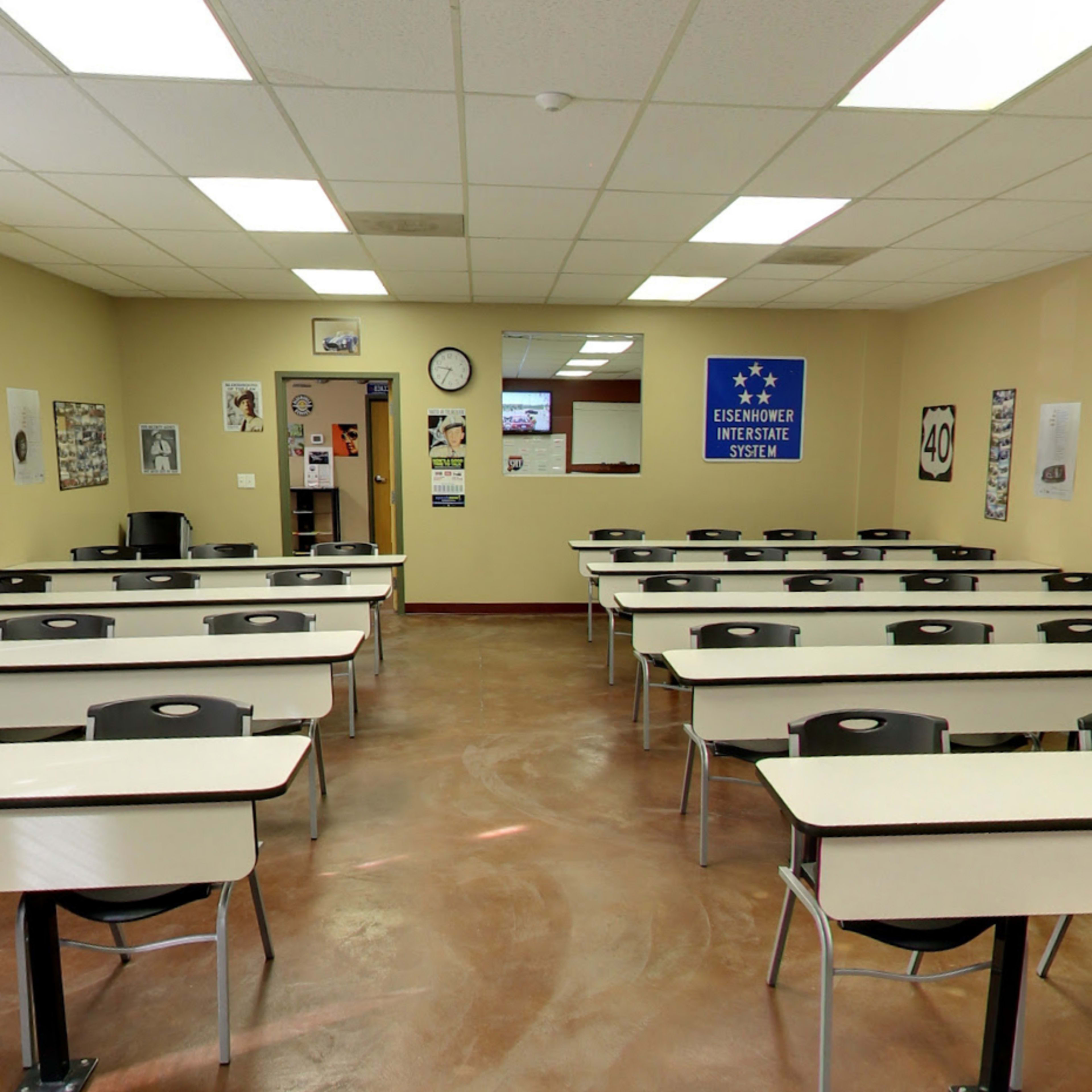 A classroom with rows of tables and chairs, posters on the walls, and a door at the back.