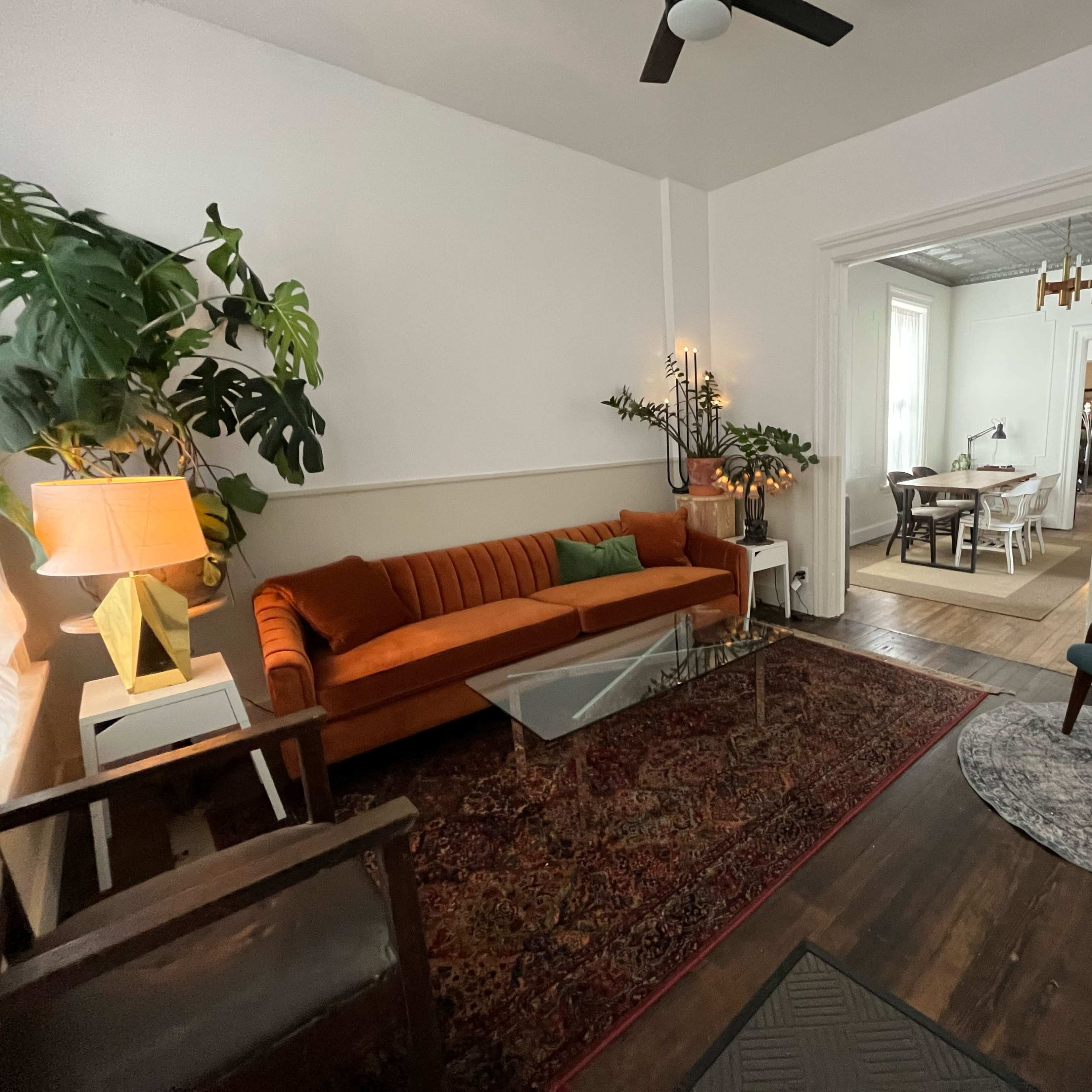 A cozy living room featuring an orange sofa, a glass coffee table, and a green armchair, with plants and decorative lighting adding to the ambiance.
