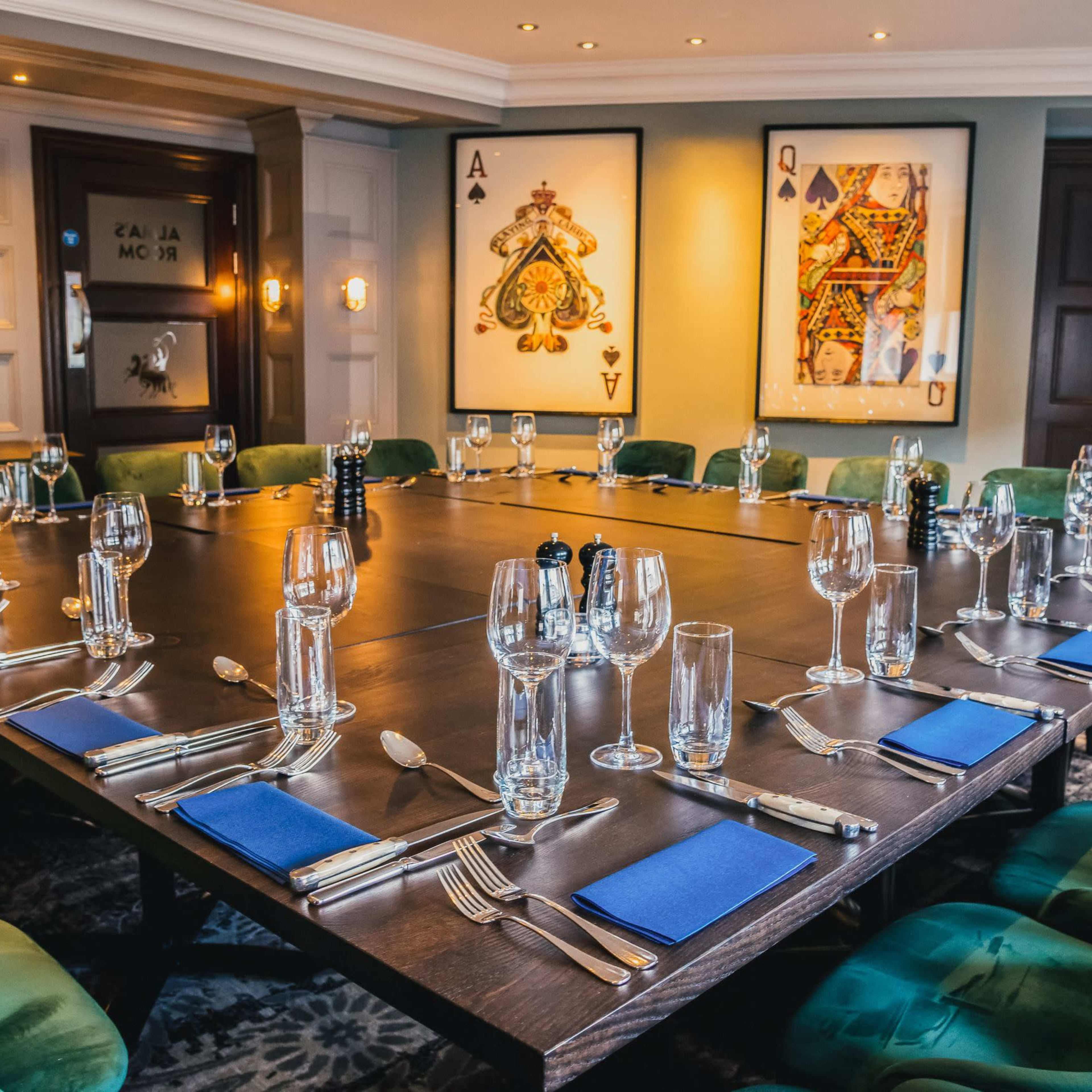 A large dining table is set with glassware, silverware, and blue napkins in a well-decorated room featuring playing card artwork.