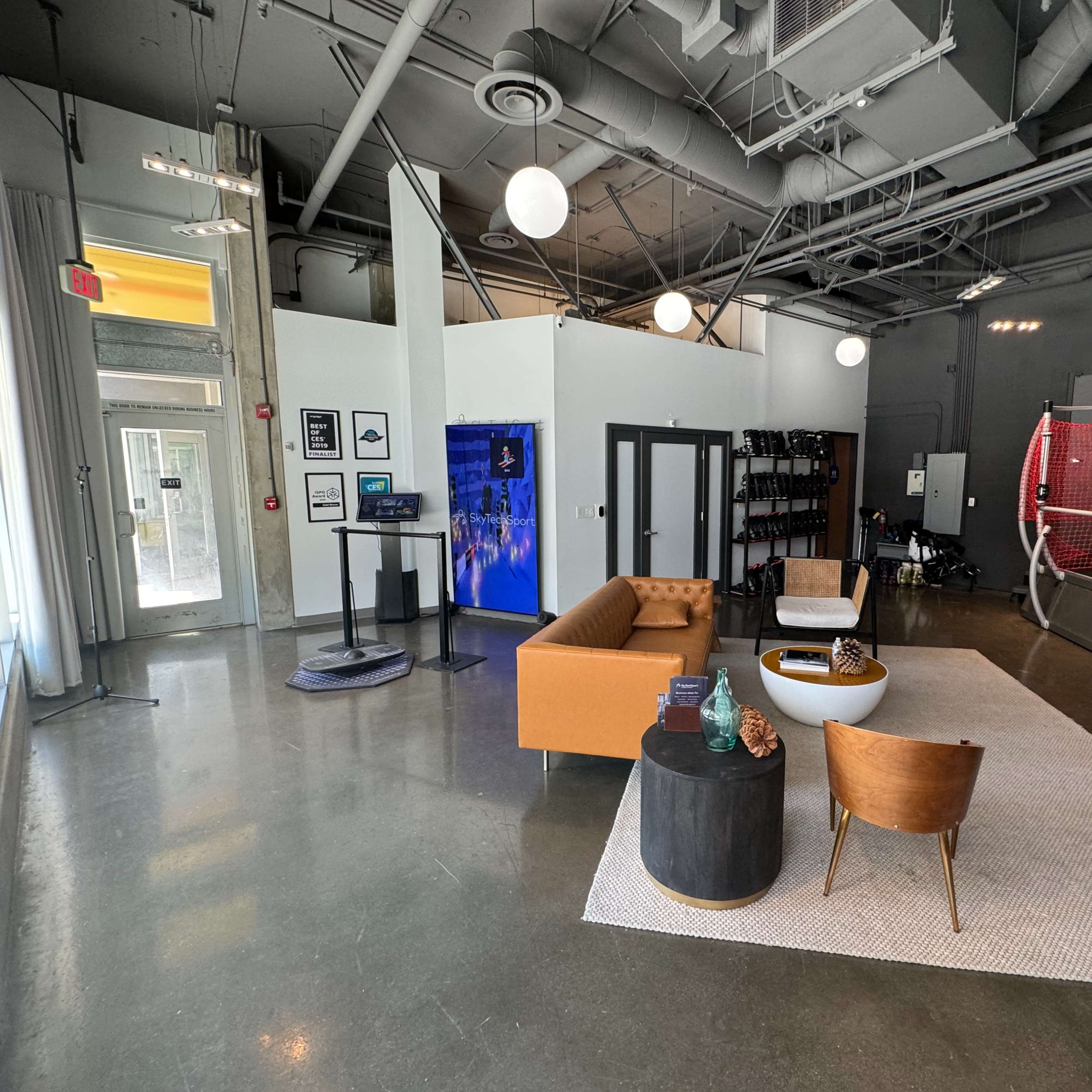 A modern open space with a seating area featuring a brown sofa and black coffee table, alongside a wall-mounted display and a sports court area in the background.