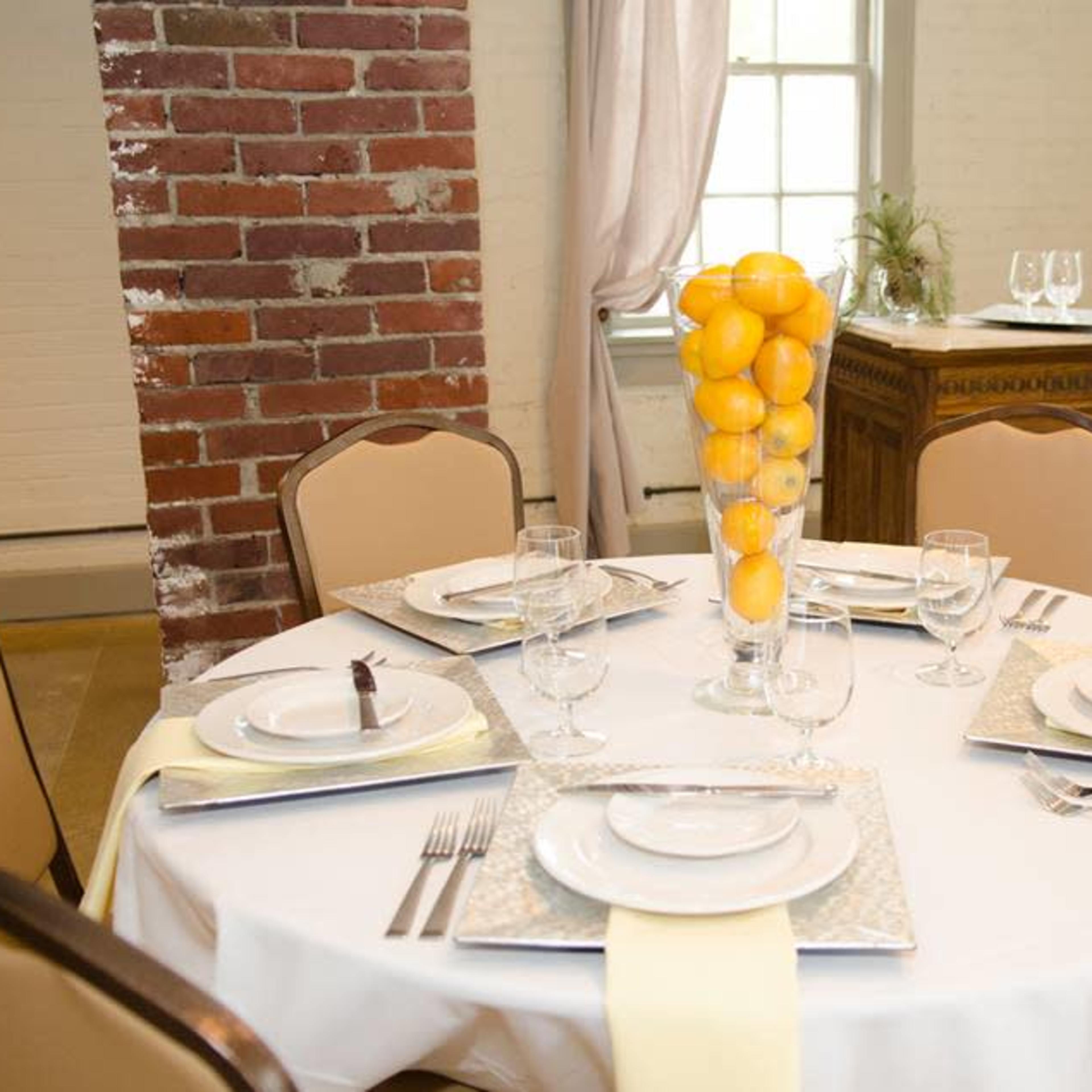 A round dining table is set with white plates, glassware, and a centerpiece of lemons in a clear vase, surrounded by beige chairs in a well-lit room with brick walls.
