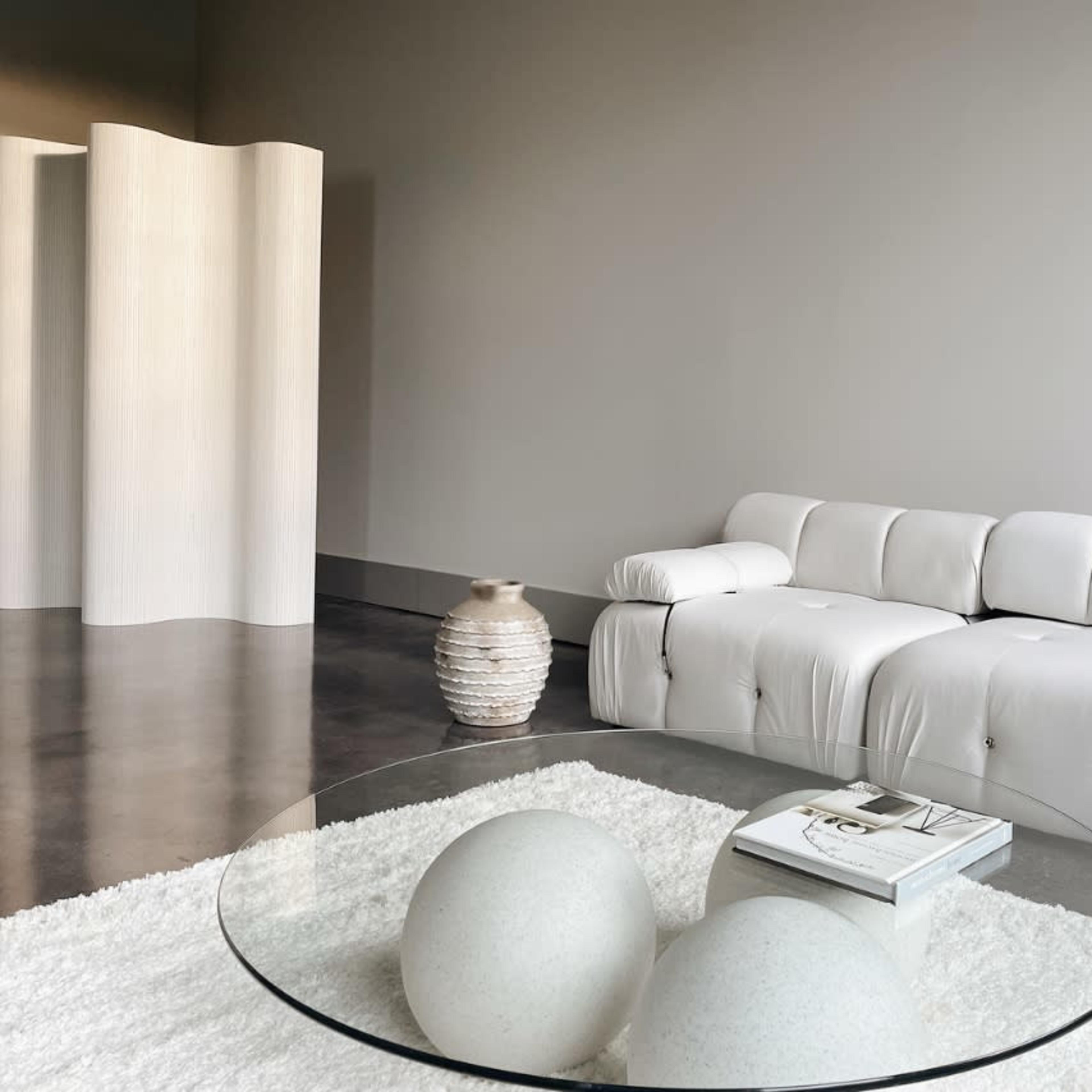 A modern, minimalist living space features a white sectional sofa, a circular glass coffee table with two spherical decor pieces, and a textured vase against a neutral wall.
