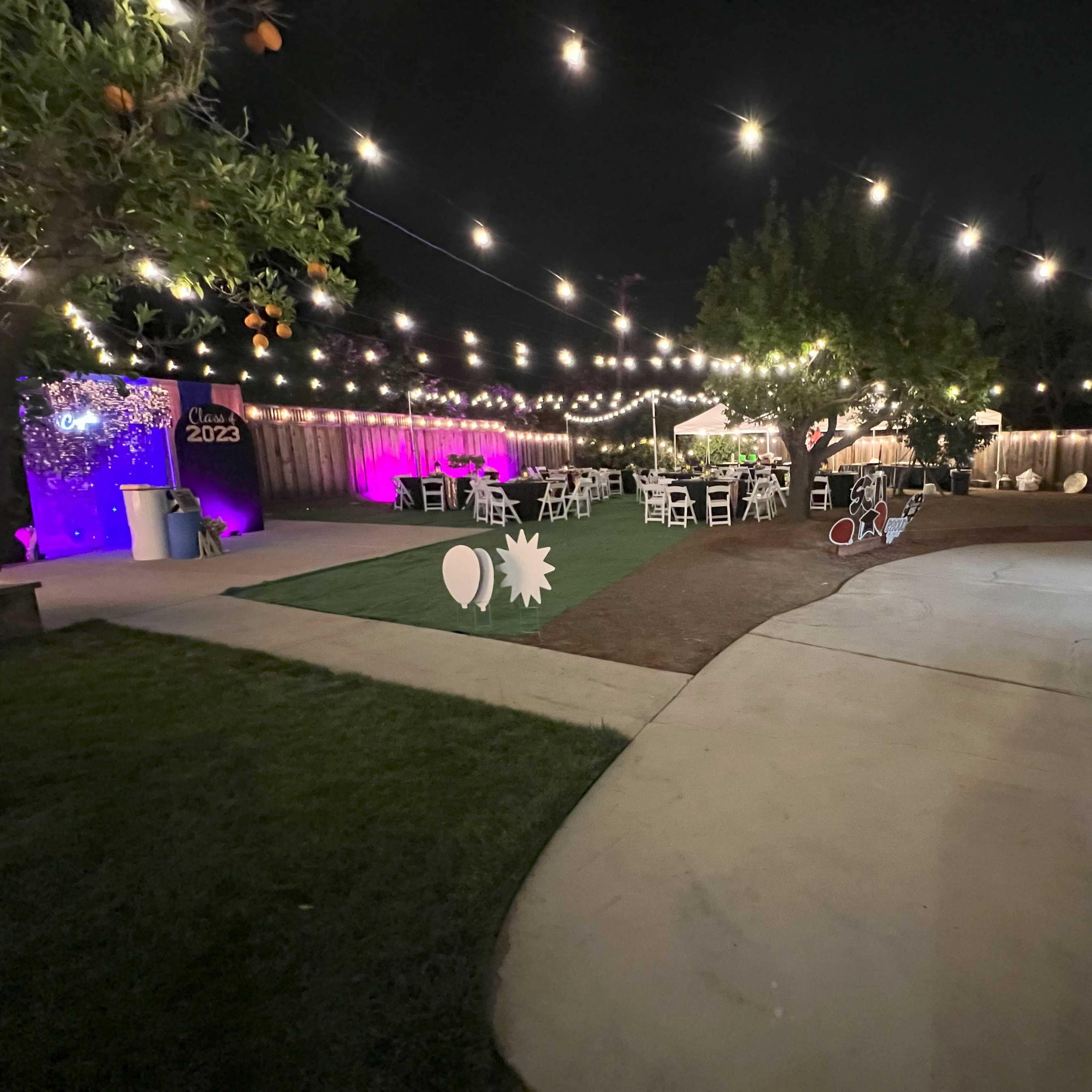 A backyard decorated for a nighttime event, featuring string lights overhead, white chairs arranged at tables, and a lush green lawn surrounded by trees.