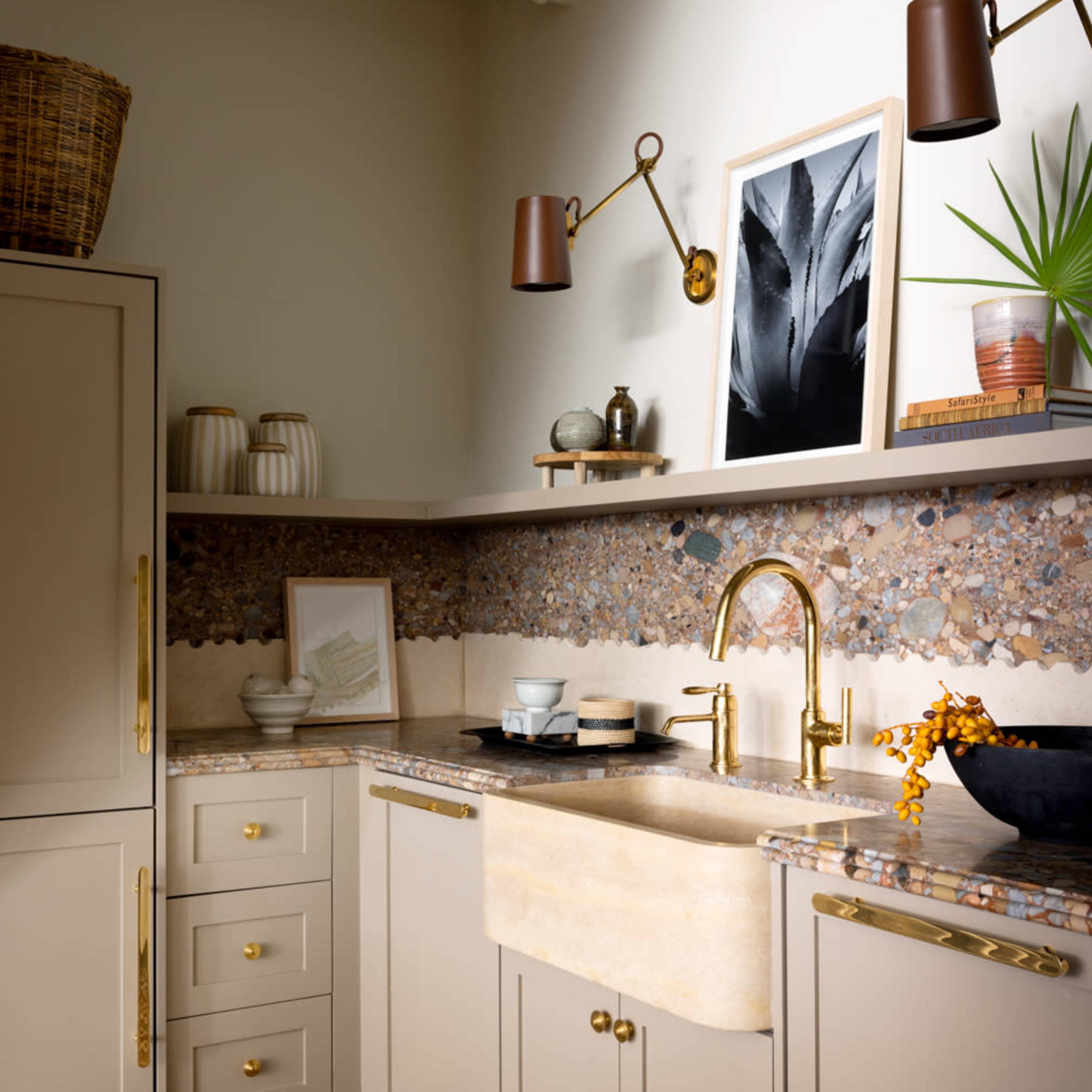 A modern kitchen features a farmhouse sink, wooden shelves with decorative items, and a textured backsplash made of various stones.