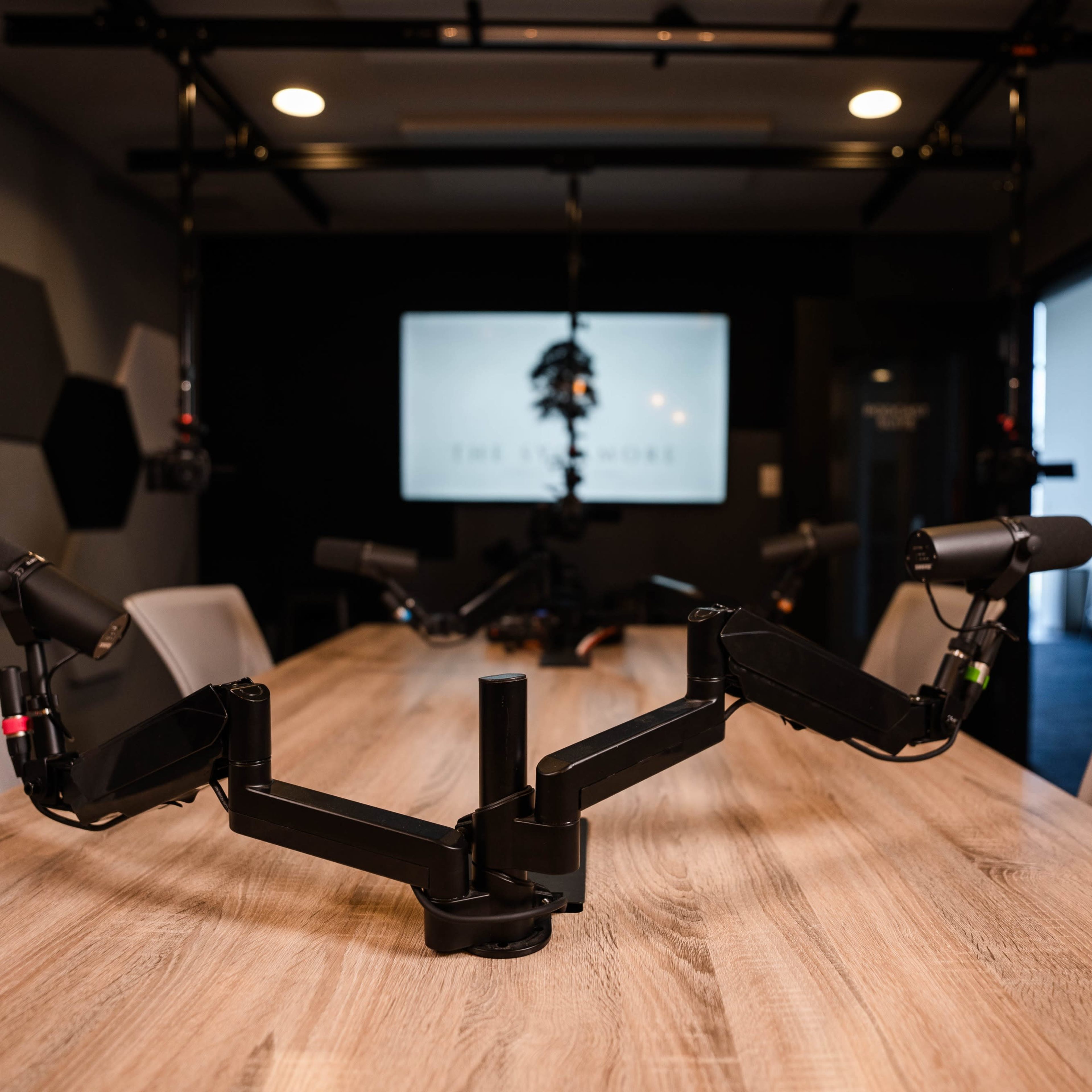 A modern conference room setup with a wooden table featuring multiple microphone arms positioned in front of a screen displaying a logo.