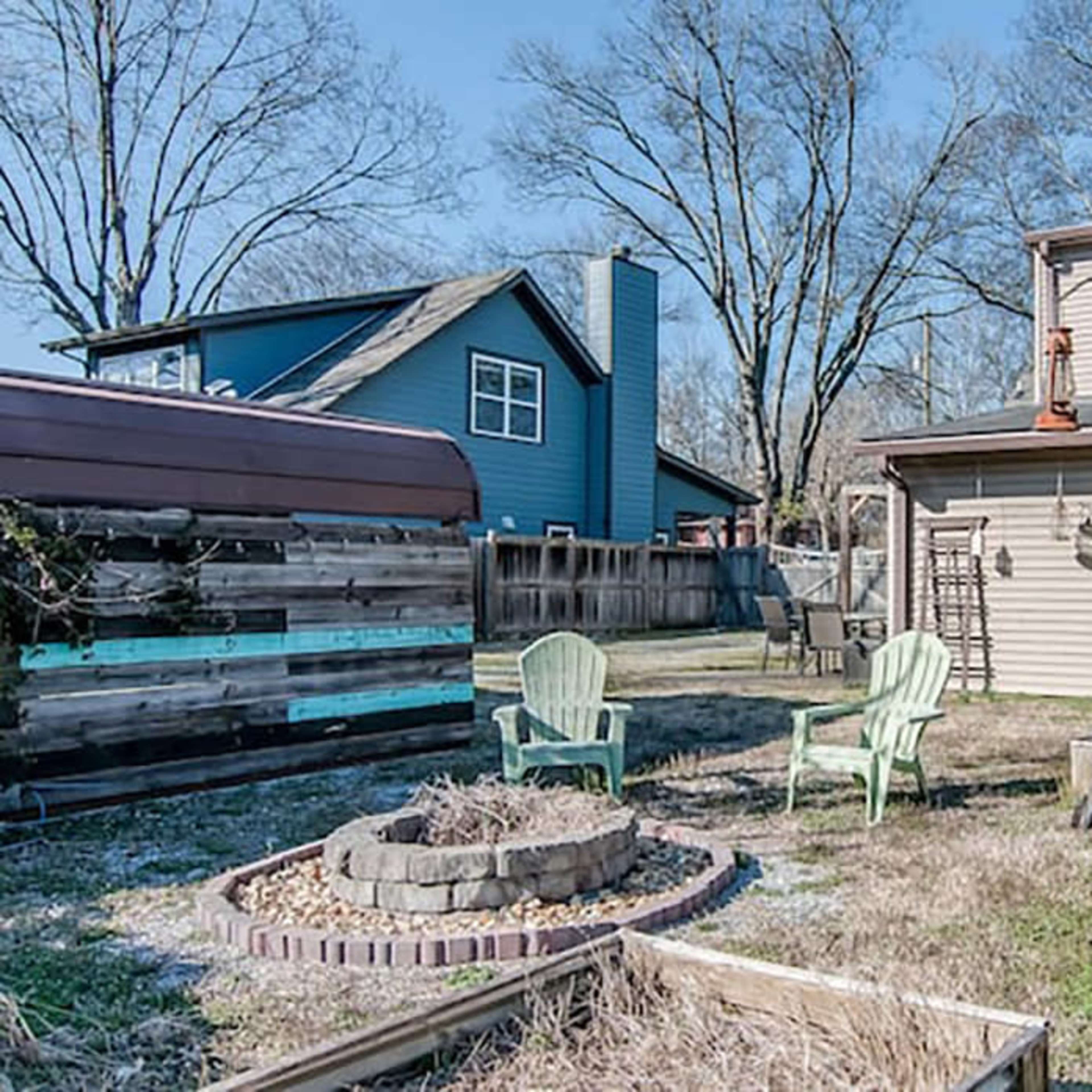 A backyard with a fire pit surrounded by two green chairs, a swing set, and a blue house in the background.