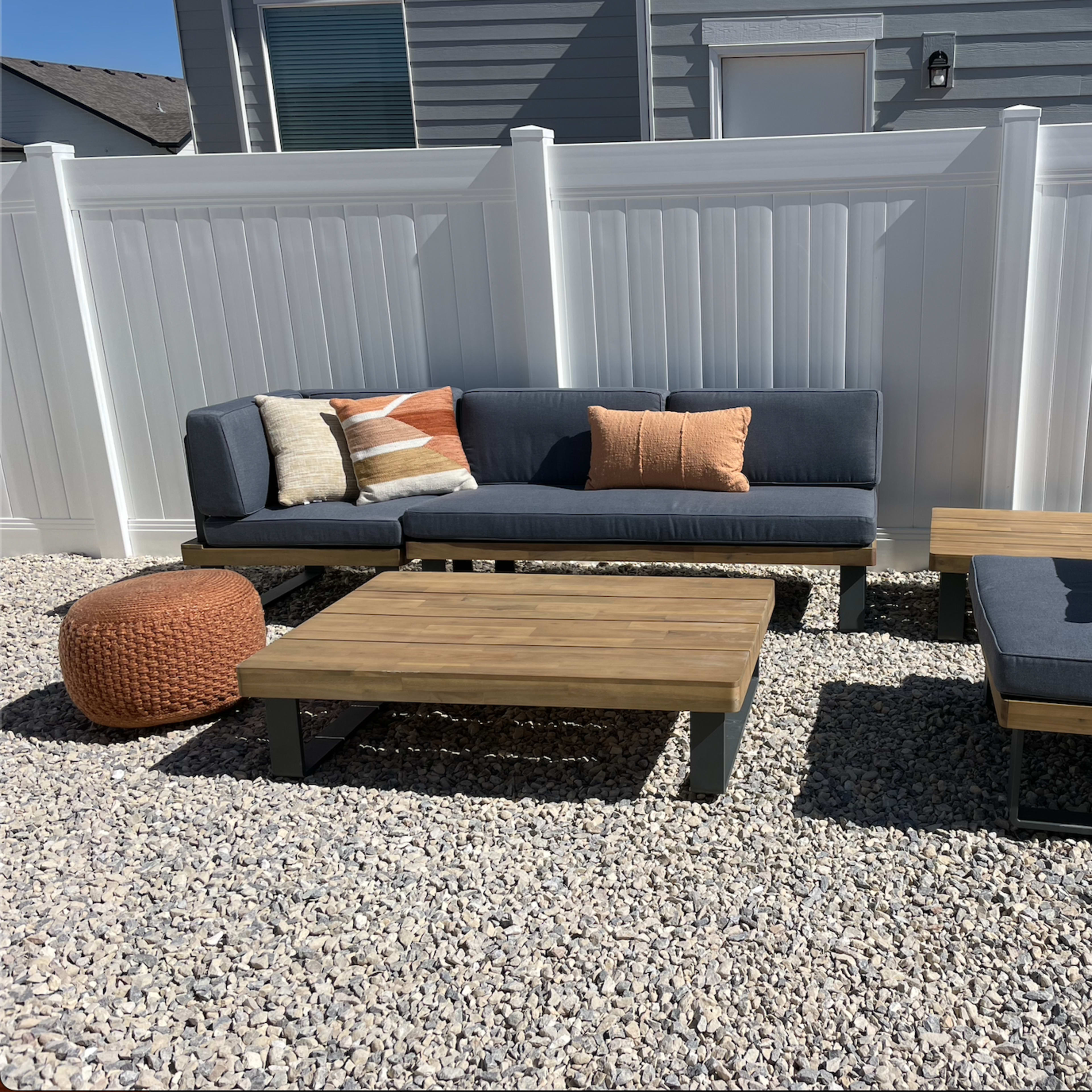 A modern outdoor seating area features a sectional sofa, a single chair, and a coffee table made of wood, all placed on gravel beneath a white vinyl fence.