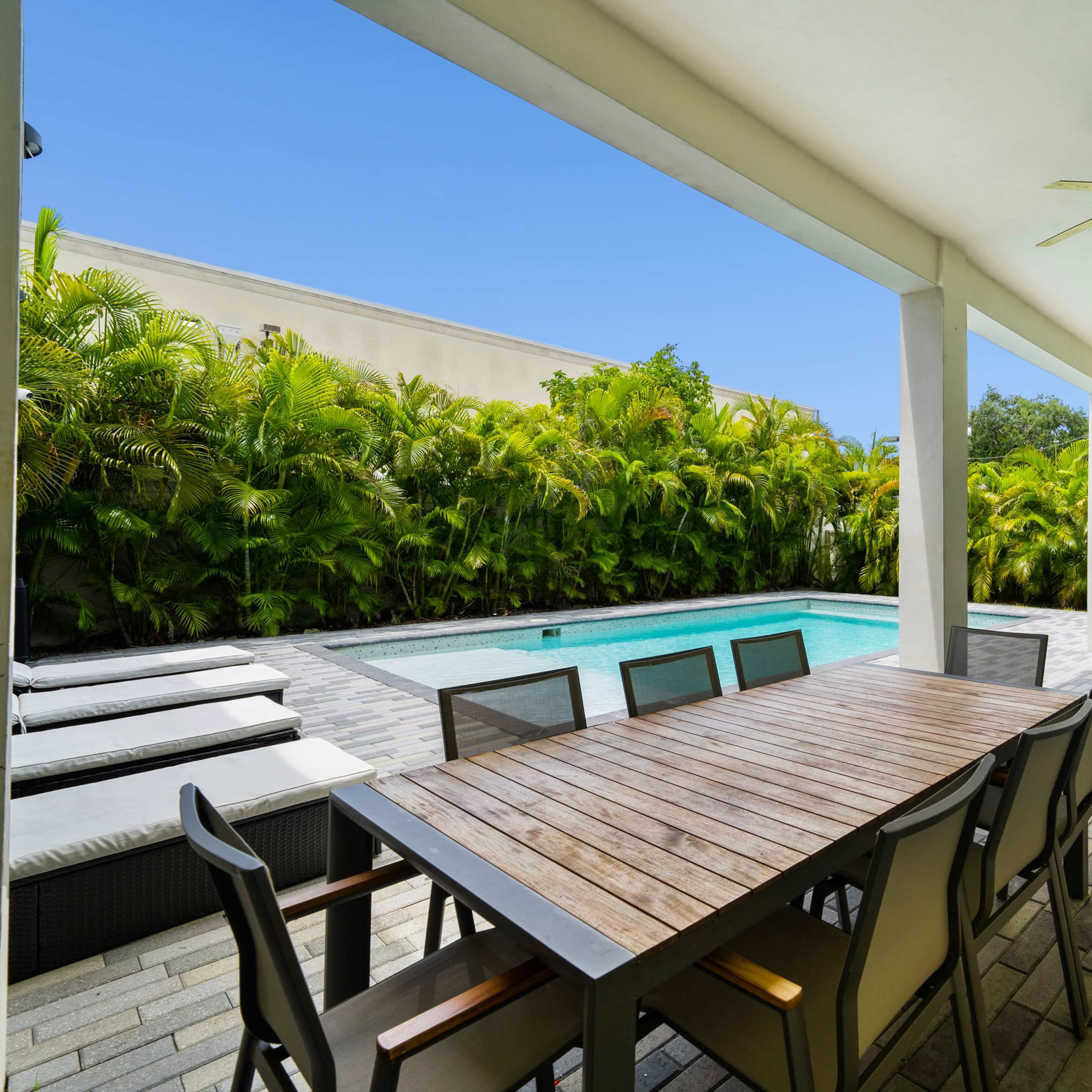 A spacious outdoor dining area with a wooden table and chairs overlooks a swimming pool surrounded by lush greenery.