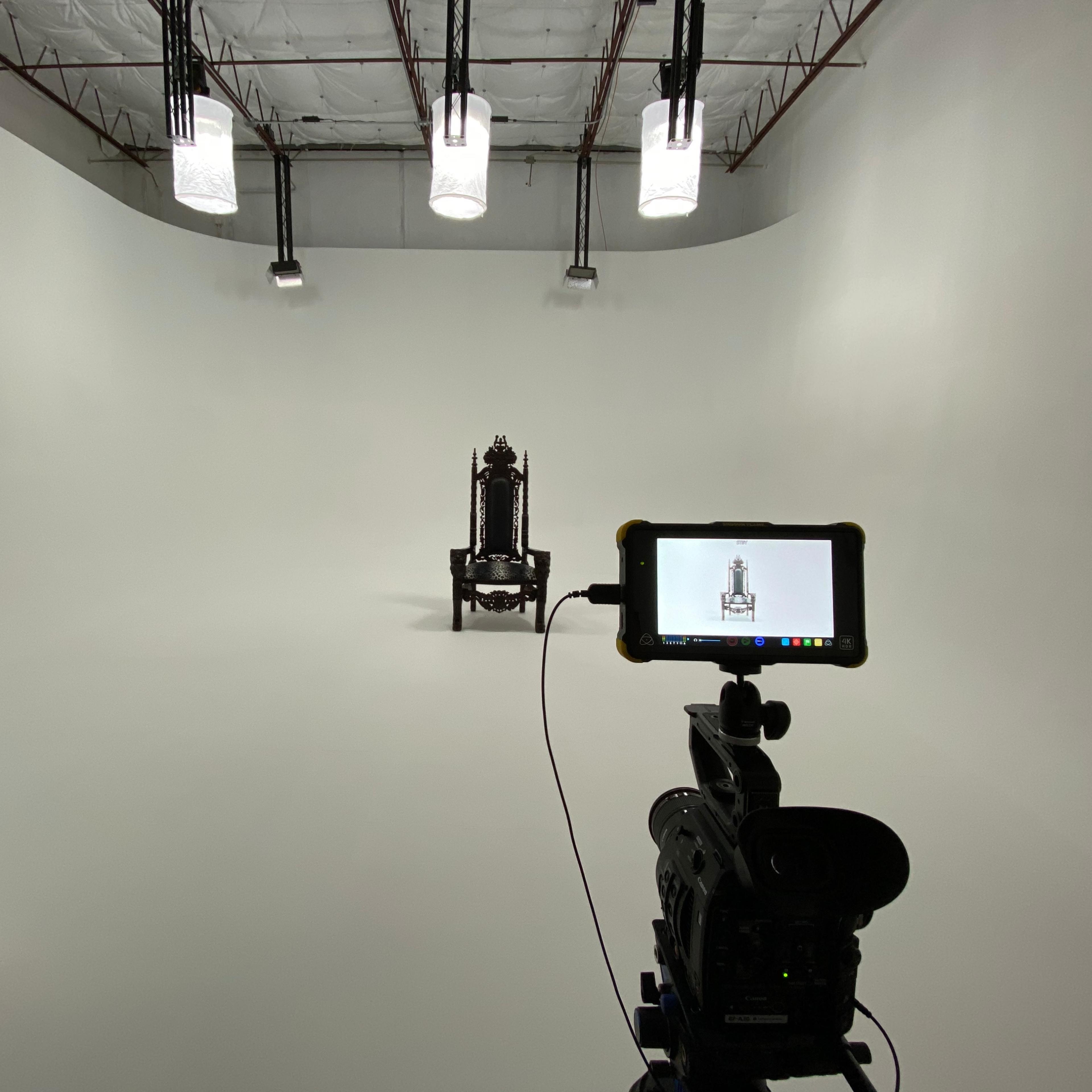 A camera is positioned in front of a vintage chair placed on a stark white background under studio lighting.