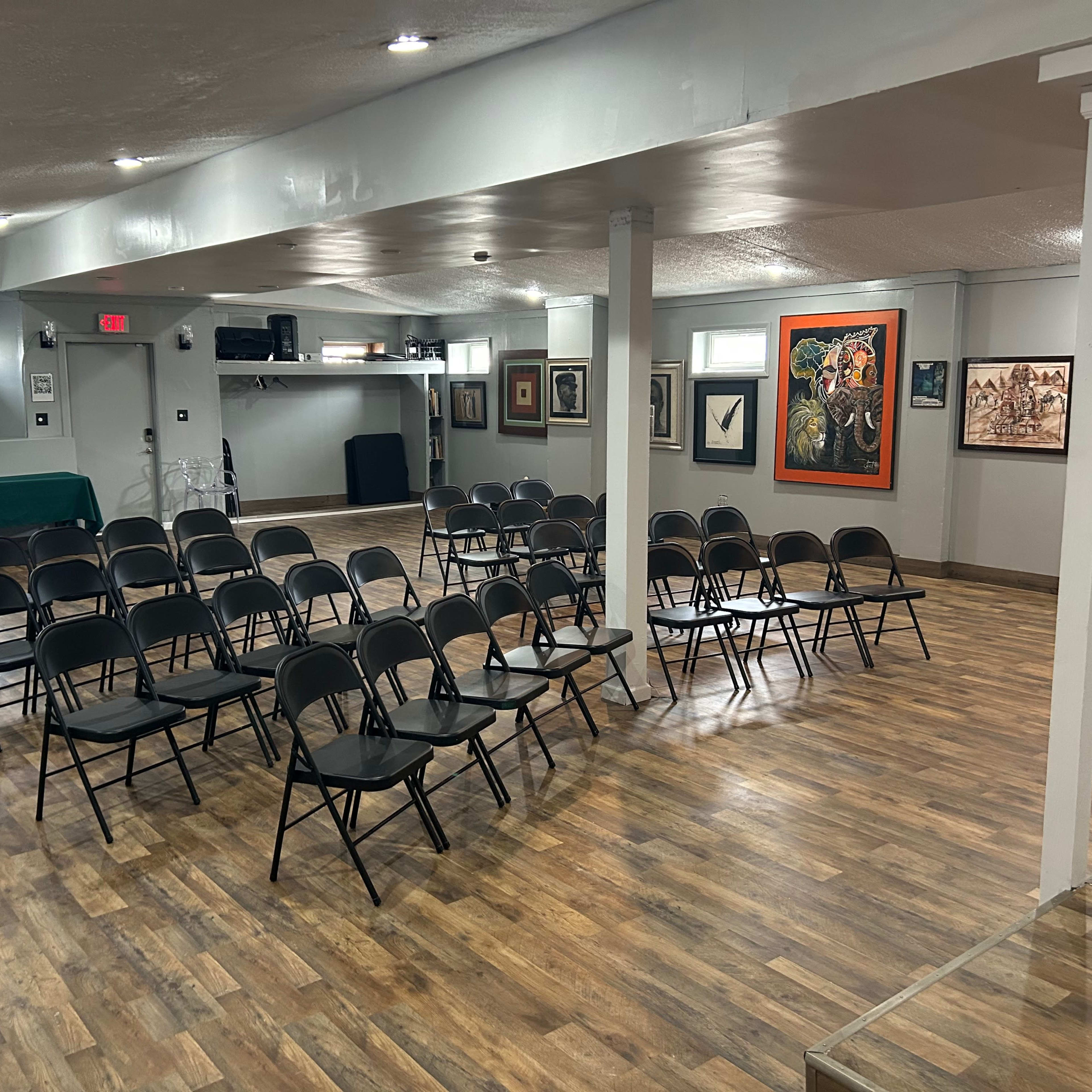 A spacious room set up with rows of black chairs facing a stage area, accompanied by various framed artworks on the walls.