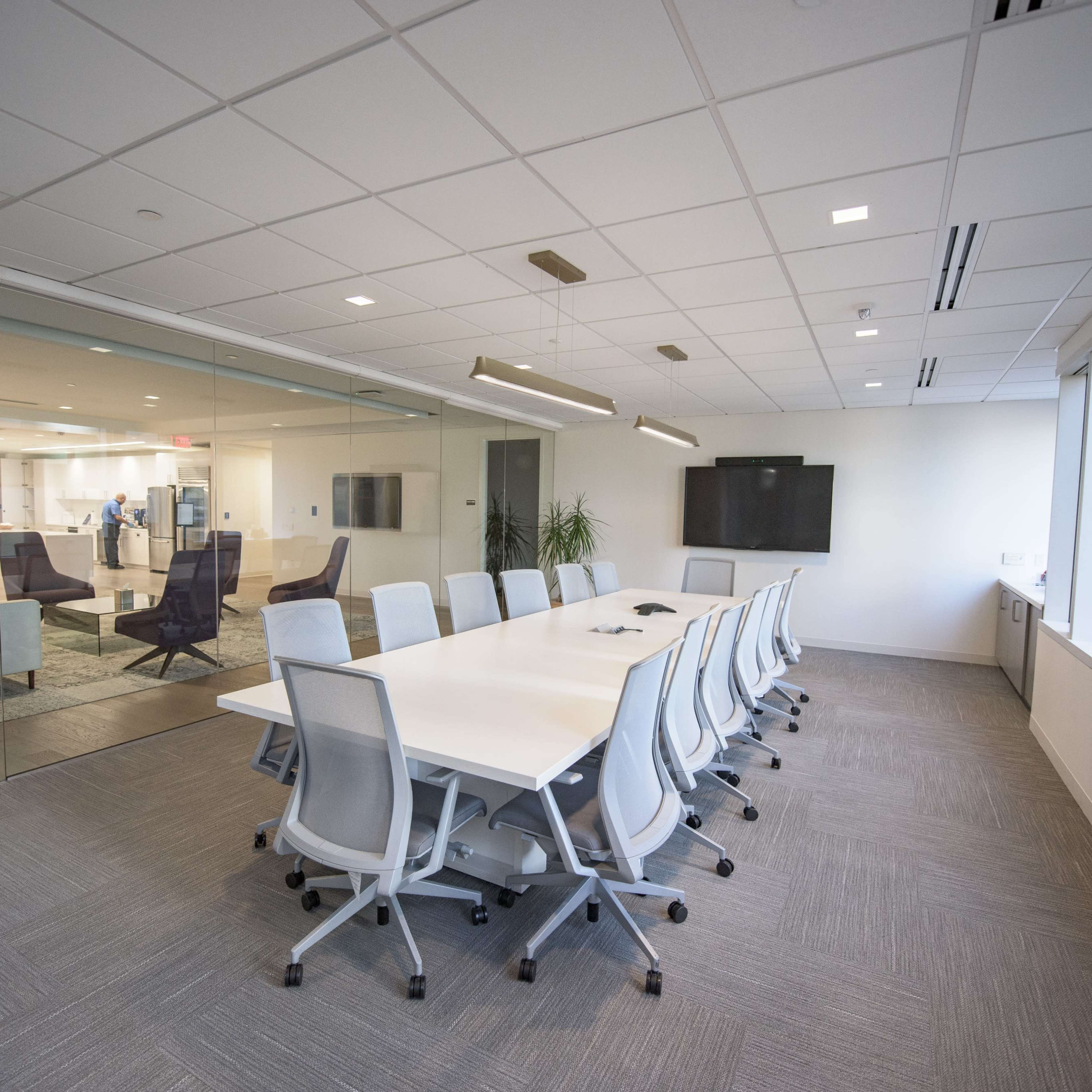 A modern conference room with a long white table surrounded by ergonomic chairs, featuring large windows and glass walls.