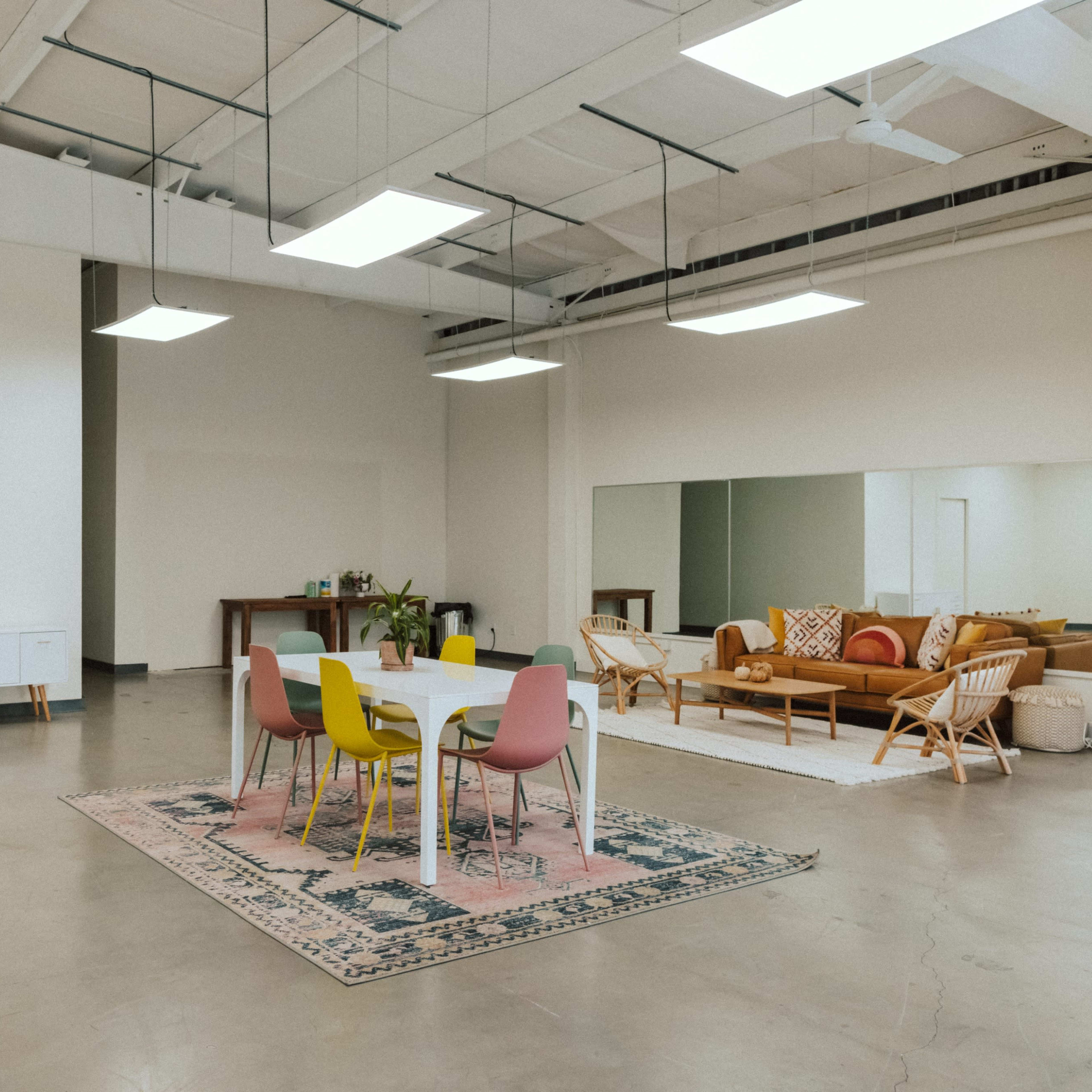 A spacious, minimalist room with a central dining table surrounded by colorful chairs, a couch, and a large mirror on one side.