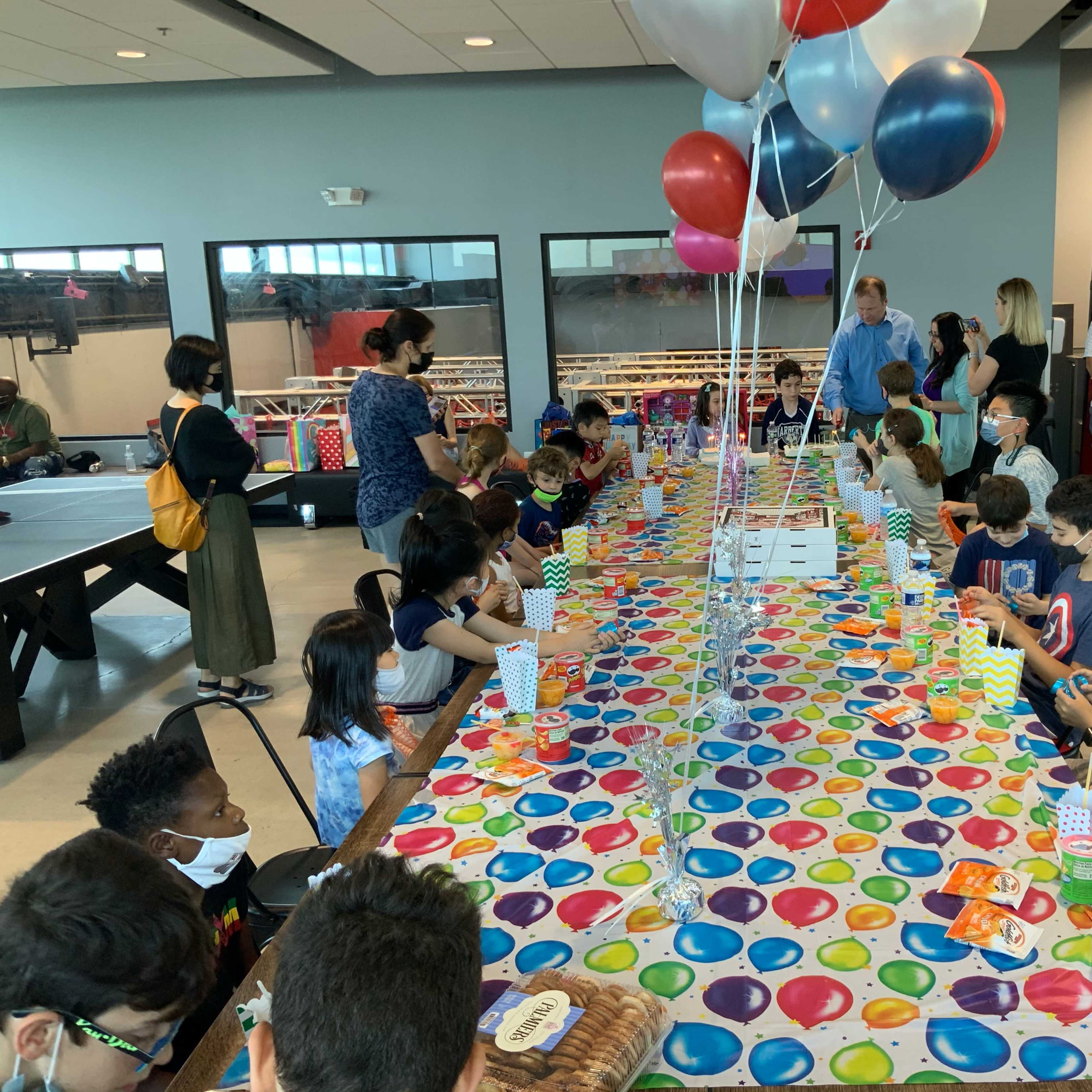 A long table covered with a colorful tablecloth is set for a gathering of children and adults, with balloons overhead and various party items arranged along the tabletop.