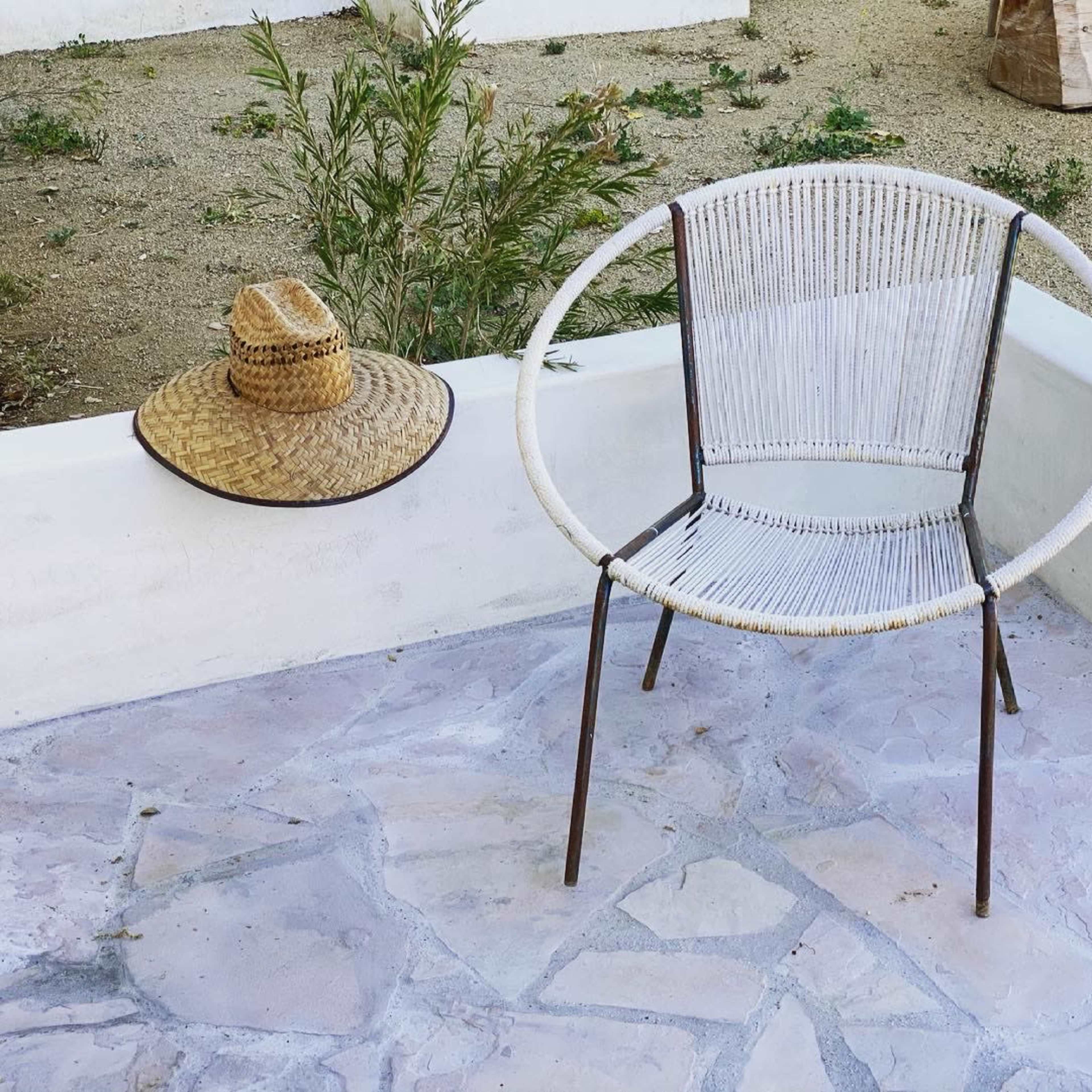 A white circular chair with a metal frame sits on a stone patio next to a straw hat and surrounded by gravel and sparse vegetation.