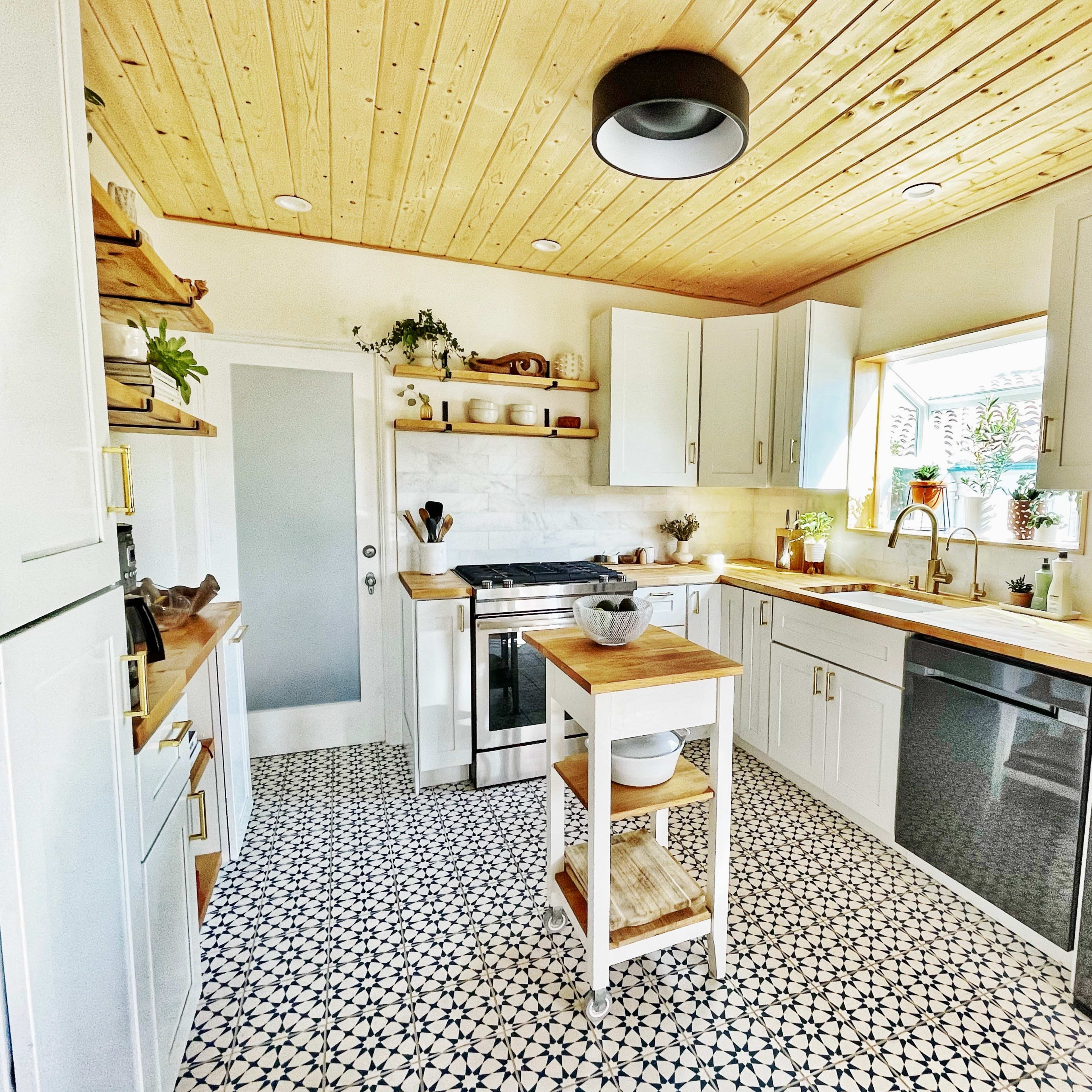 A modern kitchen features white cabinets, a butcher block island, and patterned tile flooring, illuminated by natural light from a window.