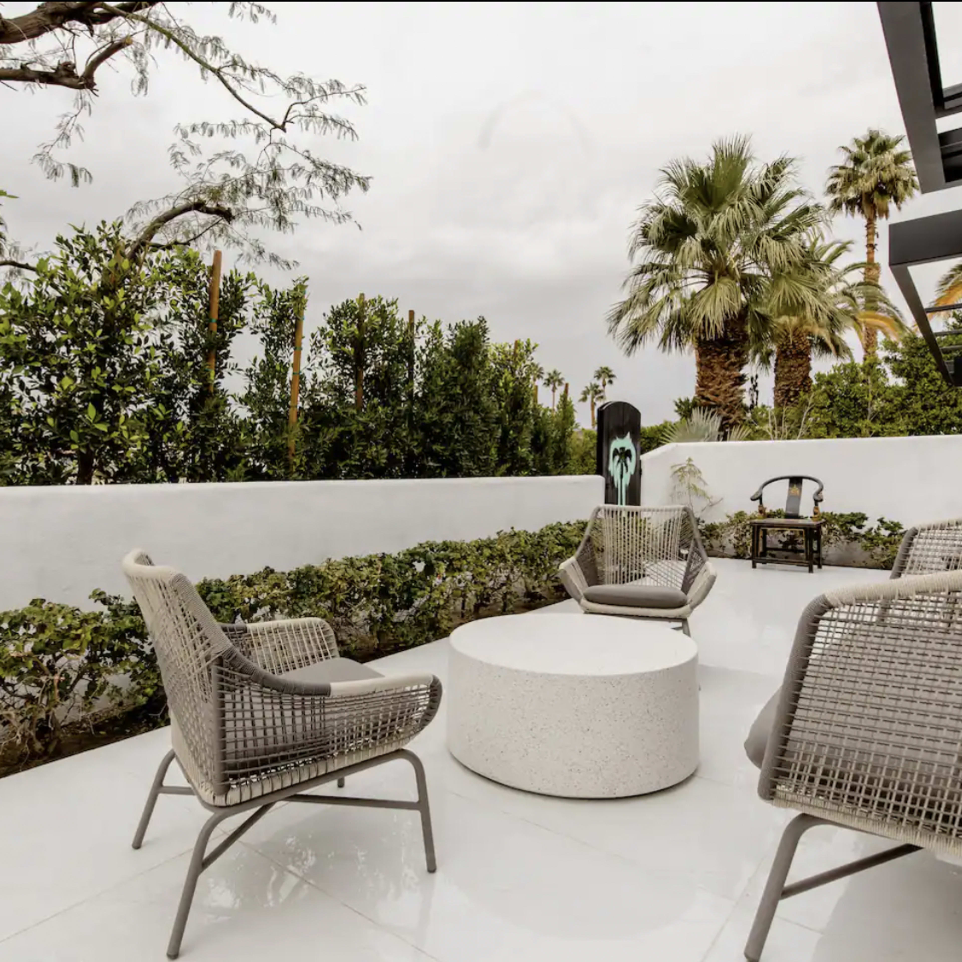 A patio features several modern chairs arranged around a round table, with lush greenery and palm trees in the background.