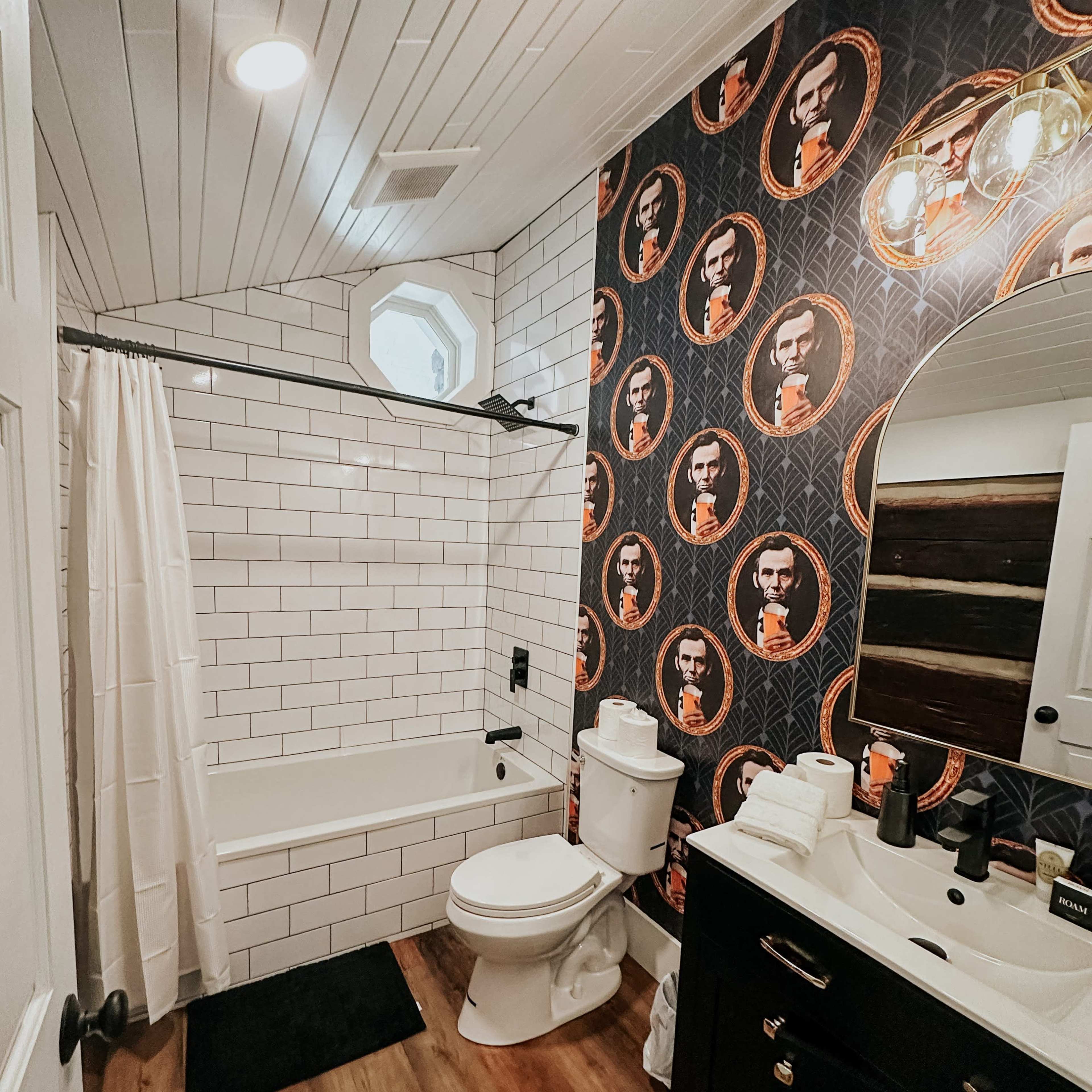 The bathroom features a white tiled bathtub and toilet, a circular mirror, and walls decorated with a patterned wallpaper featuring historical portraits.