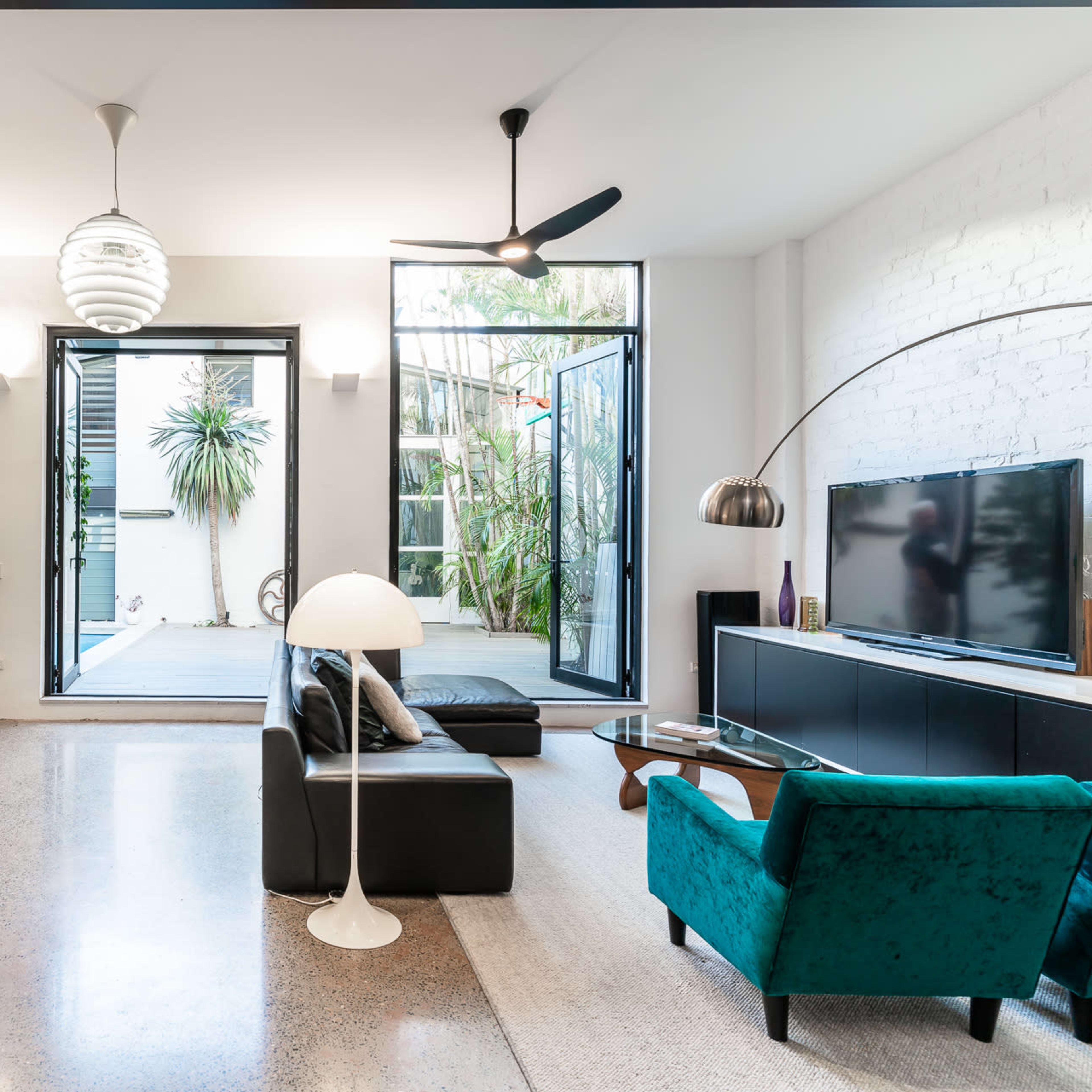 A modern living room featuring a black leather sofa, a green armchair, a large television, and glass doors opening to a patio with plants.