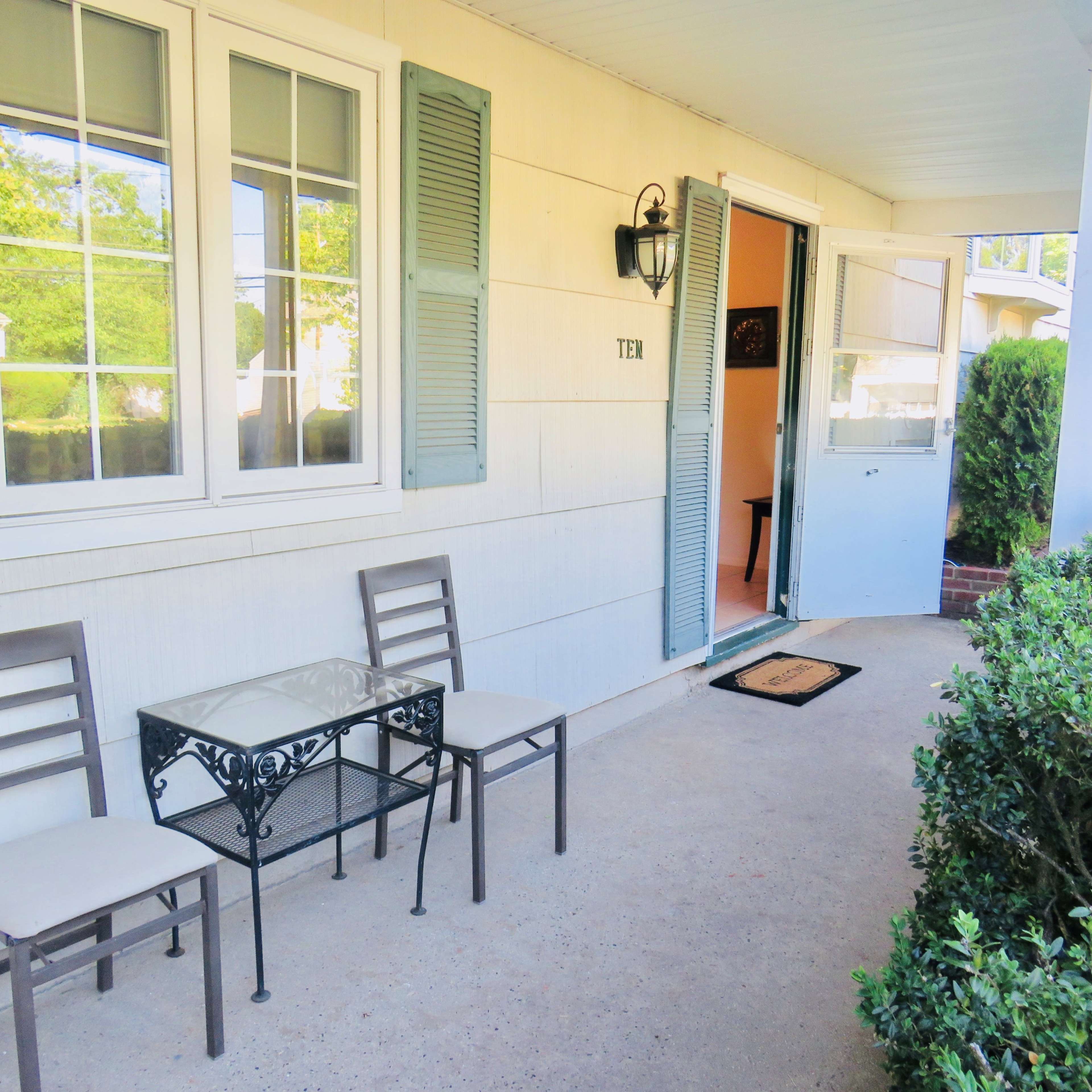 A porch with two metal chairs and a small table, flanked by green hedges and a door slightly open.