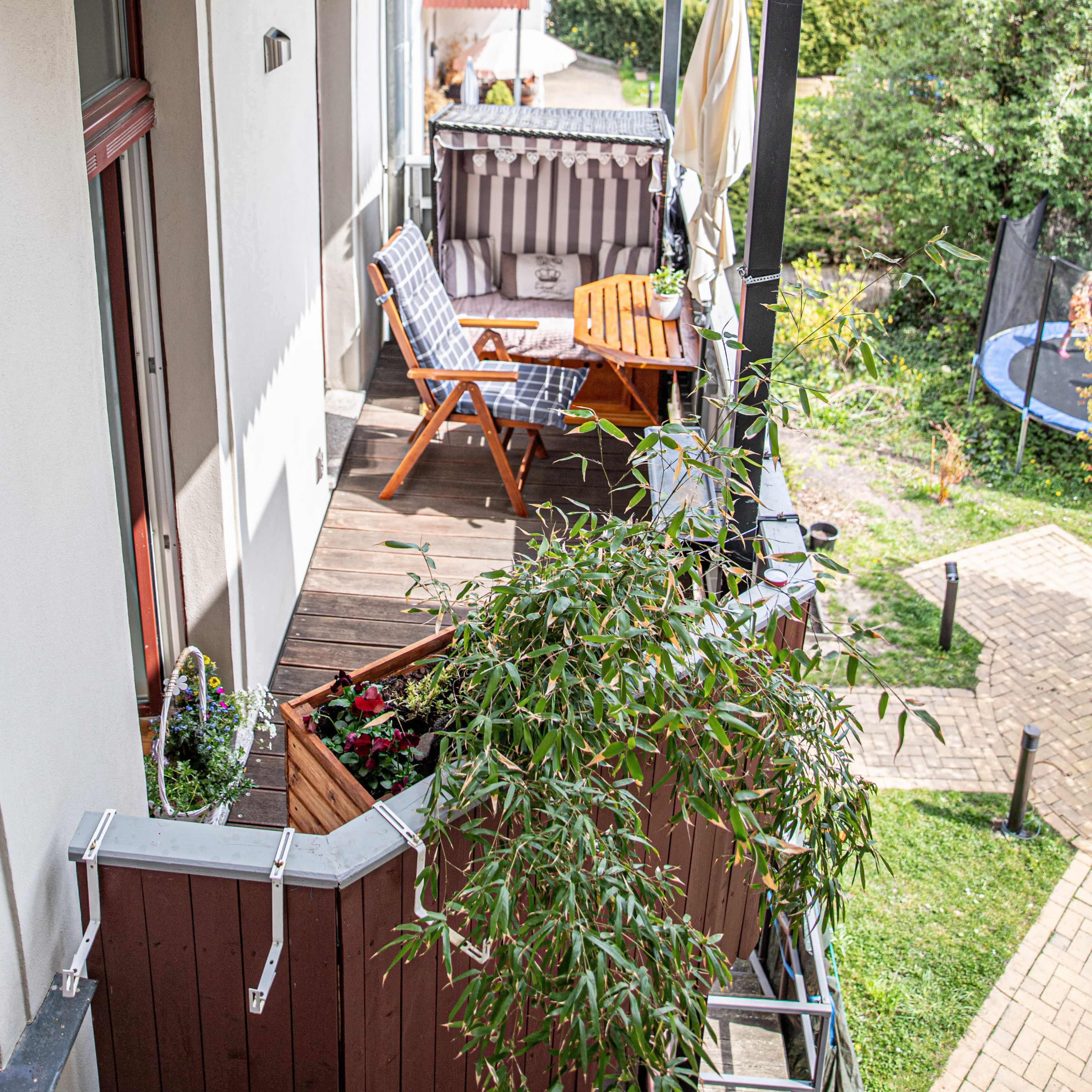 A balcony features a wooden table, a folding chair, potted plants, and a privacy screen, overlooking a green garden area.