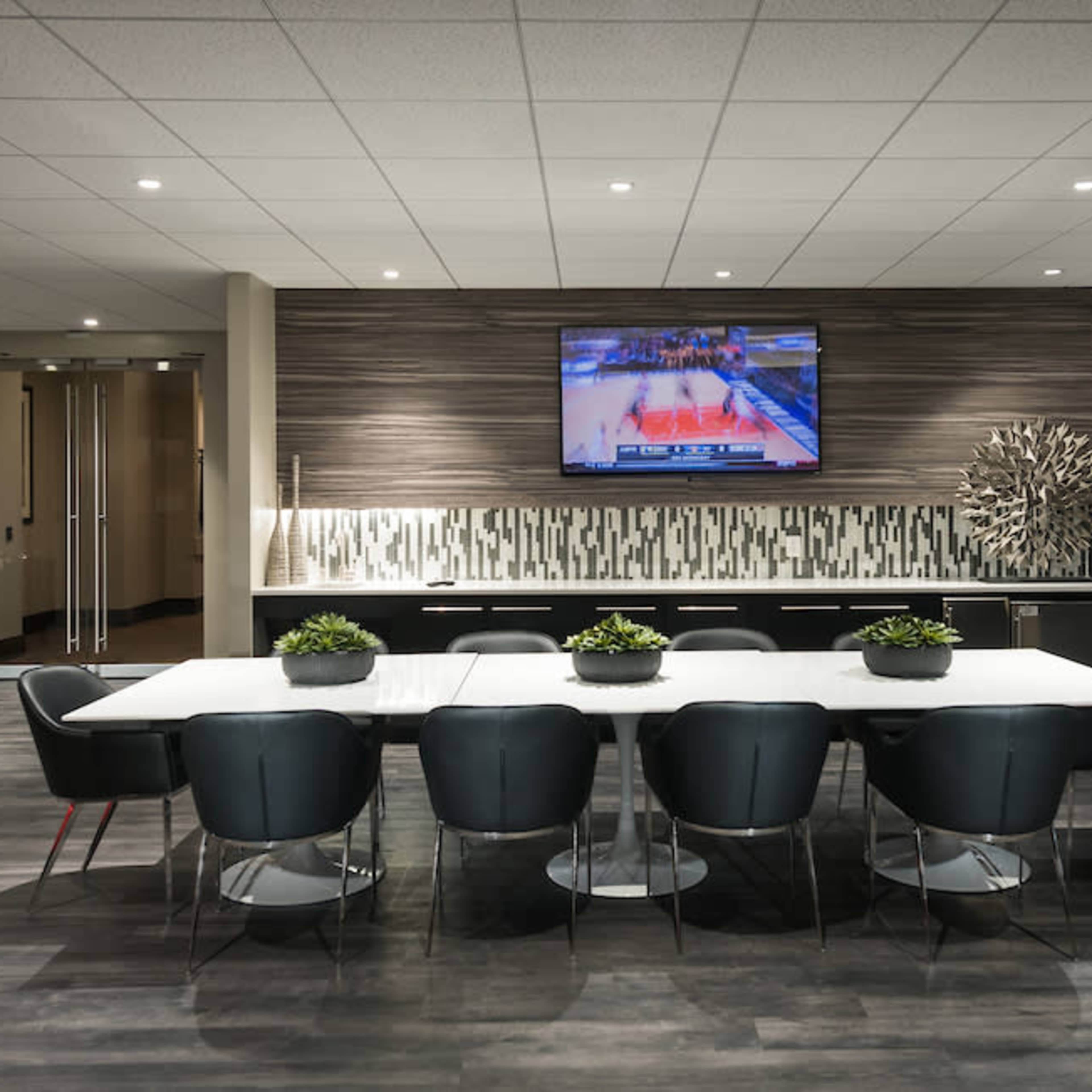 A modern conference room features a long white table surrounded by black chairs, with a television displaying a basketball game on the wall.