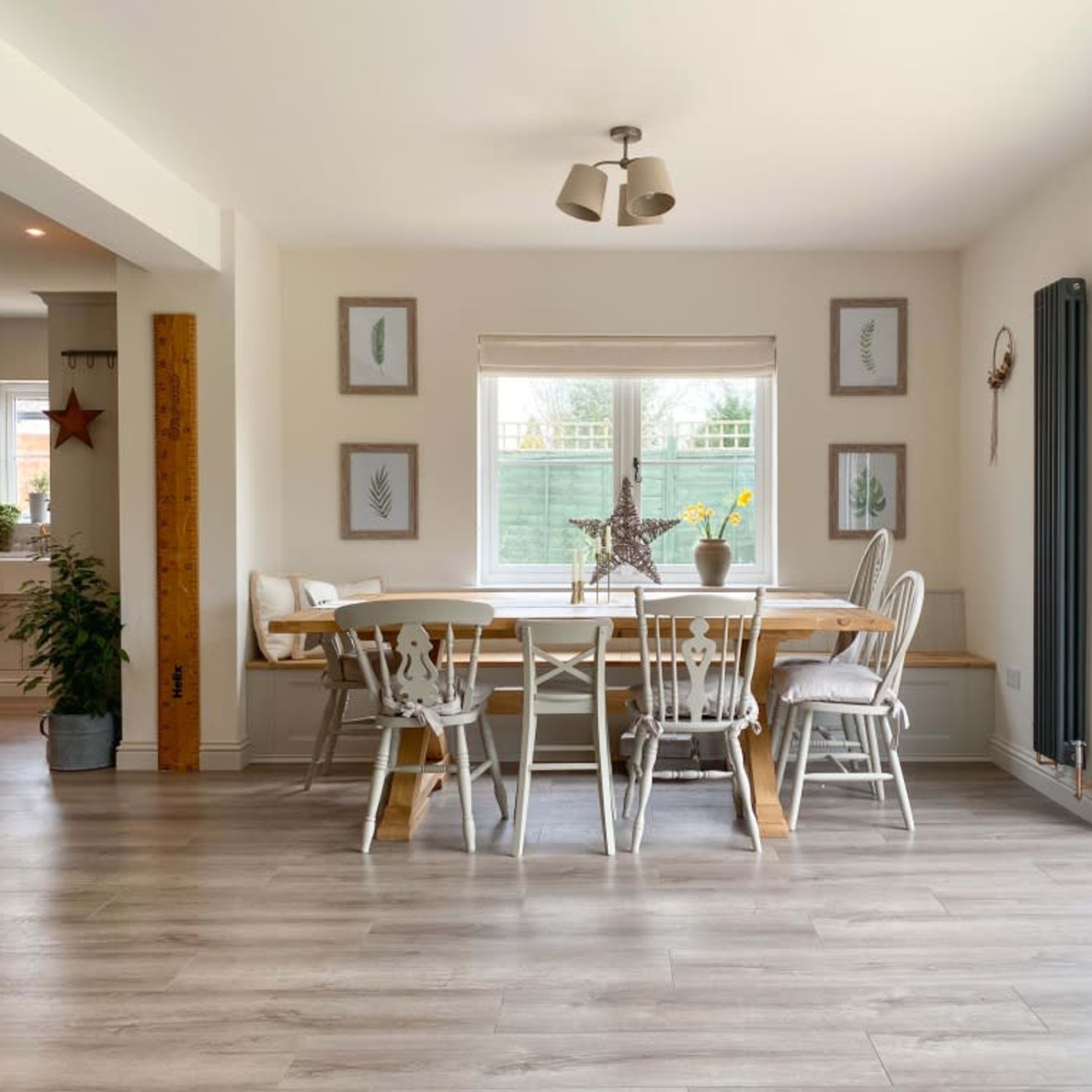 A bright dining area features a wooden table surrounded by chairs, with a window showing greenery outside and framed plant artwork on the walls.