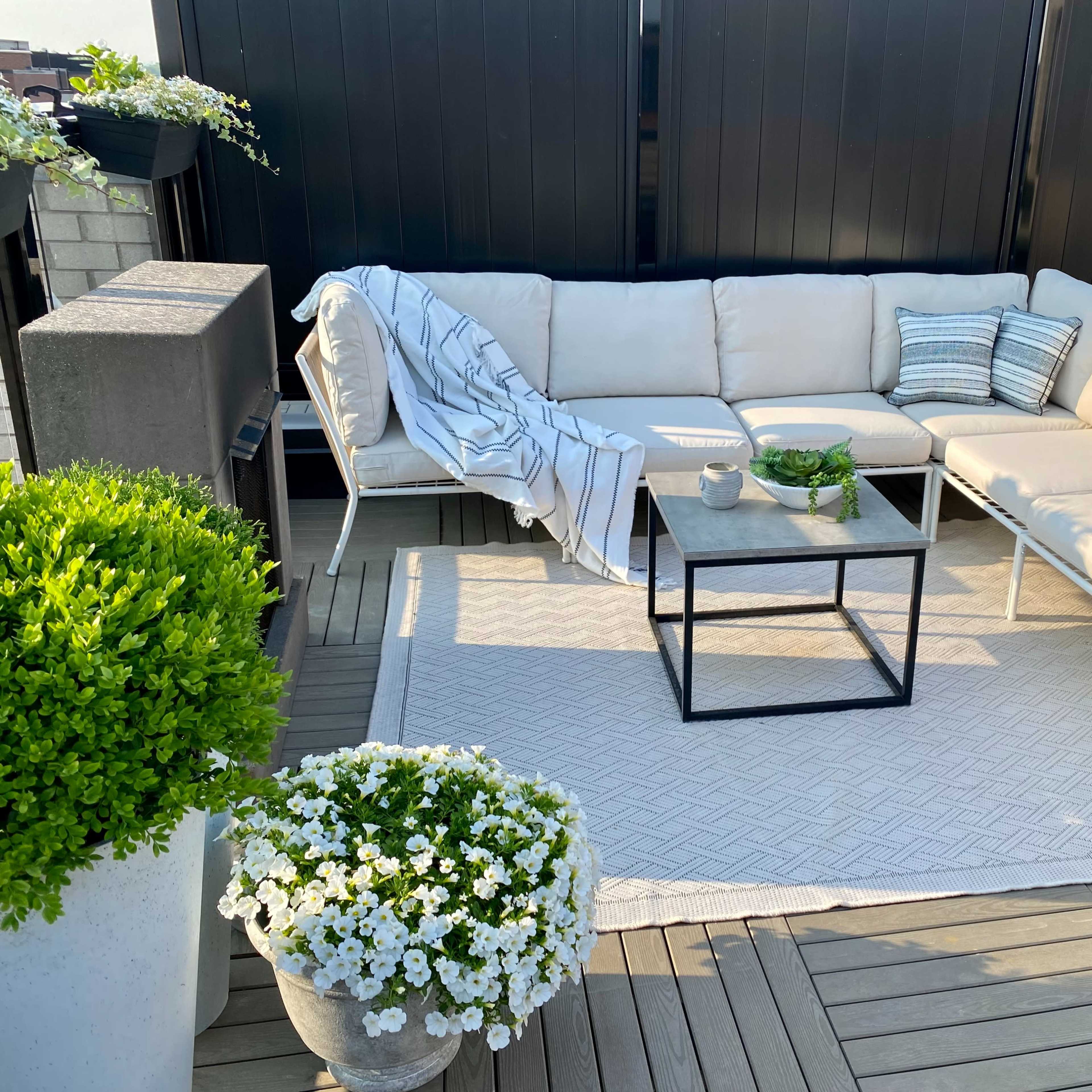 A cozy outdoor seating area features a light-colored sectional sofa, a black coffee table, and potted plants, all set on a wooden deck.