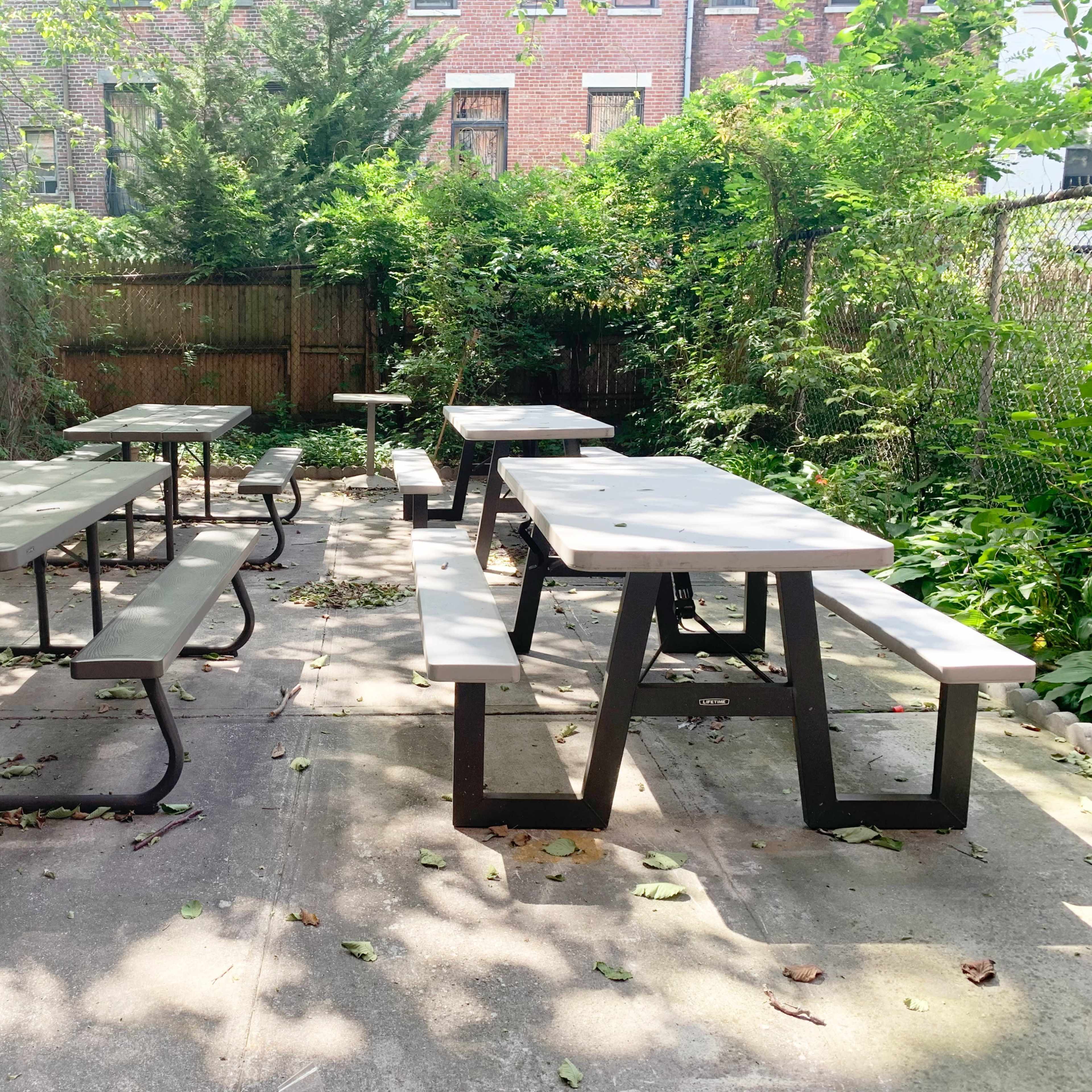 A series of metal picnic tables with benches are arranged in a garden-like outdoor space surrounded by greenery and a wooden fence.